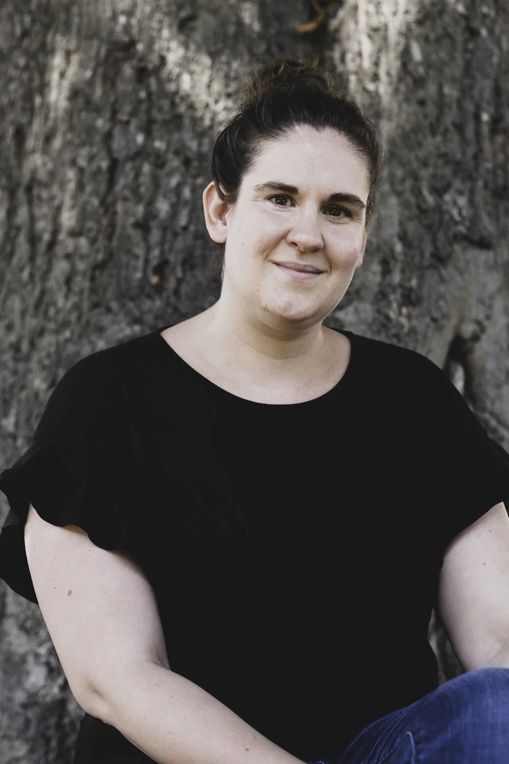 A woman with dark hair tied in a bun, wearing a black top, smiling, standing outdoors with a large tree trunk in the background.