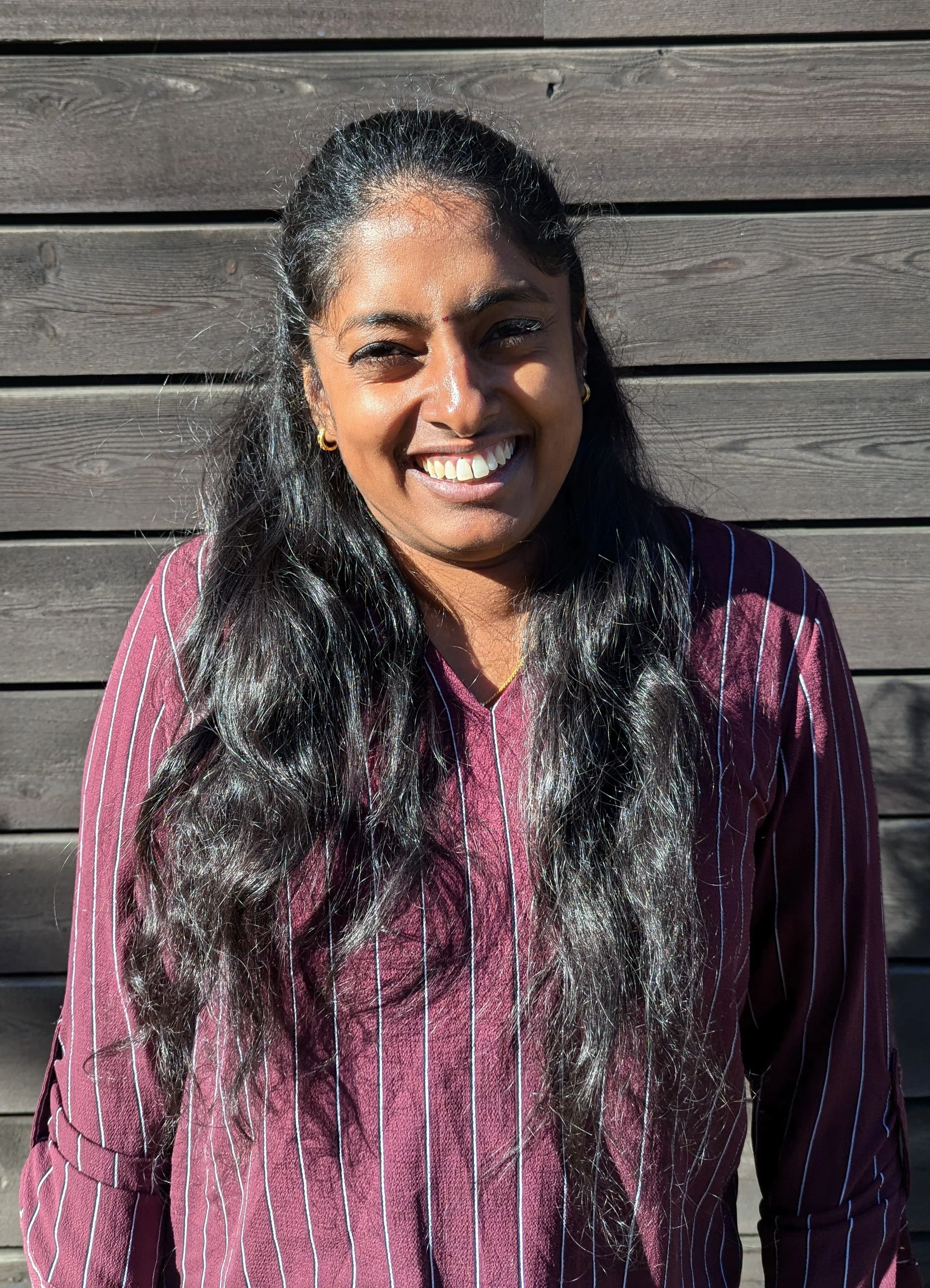 A woman with long black hair smiling outdoors in front of a wooden wall, wearing a maroon and white striped shirt.