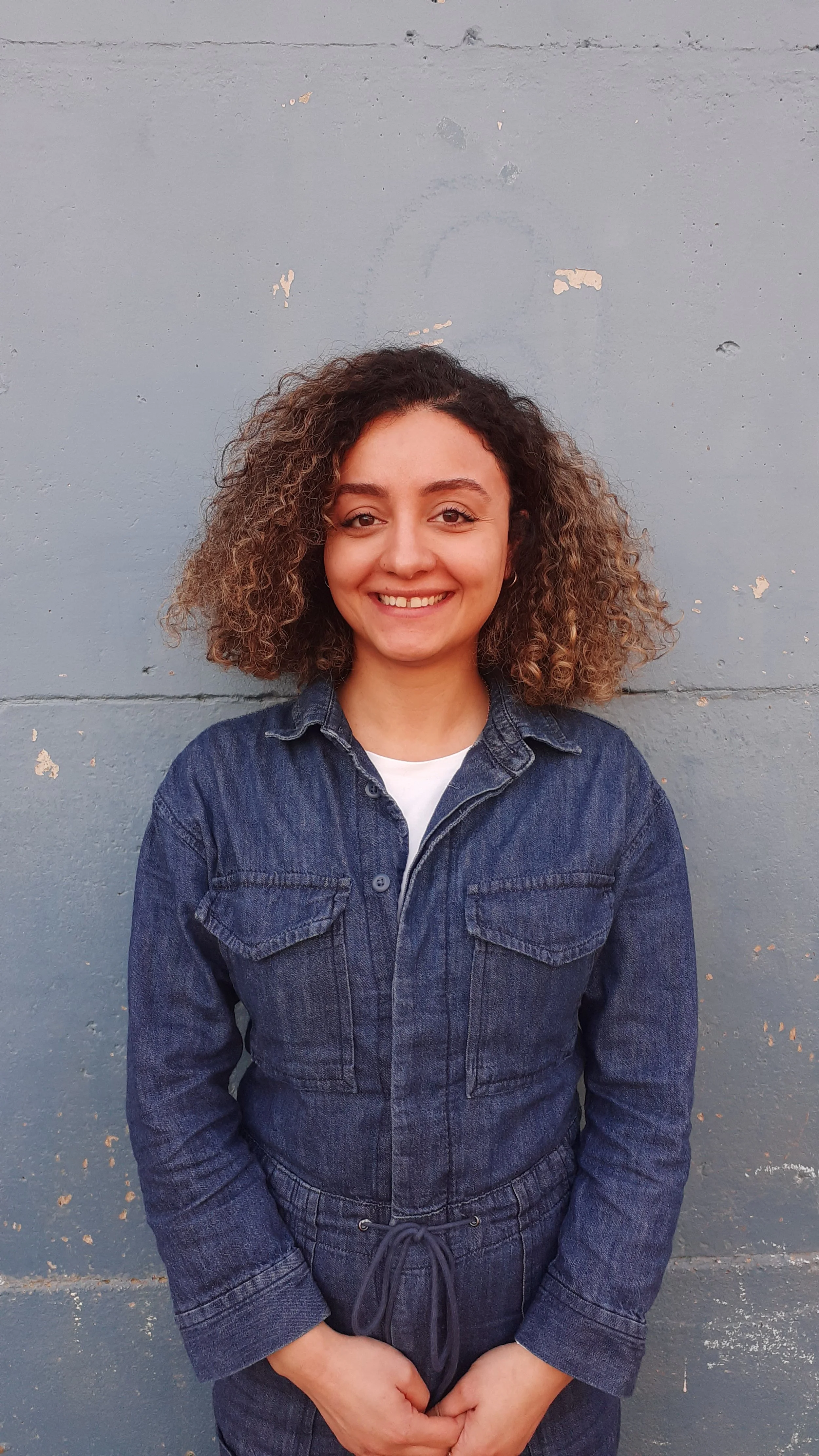 Young woman smiling, wearing a denim jumpsuit, standing against a painted concrete wall.