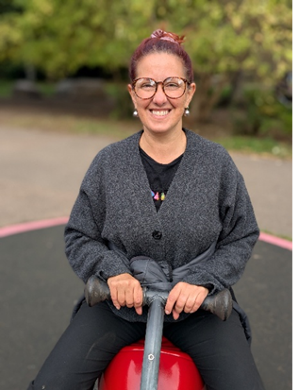 A woman with red hair, glasses, and a grey sweater sitting on a red playground toy, smiling at the camera in an outdoor park setting.