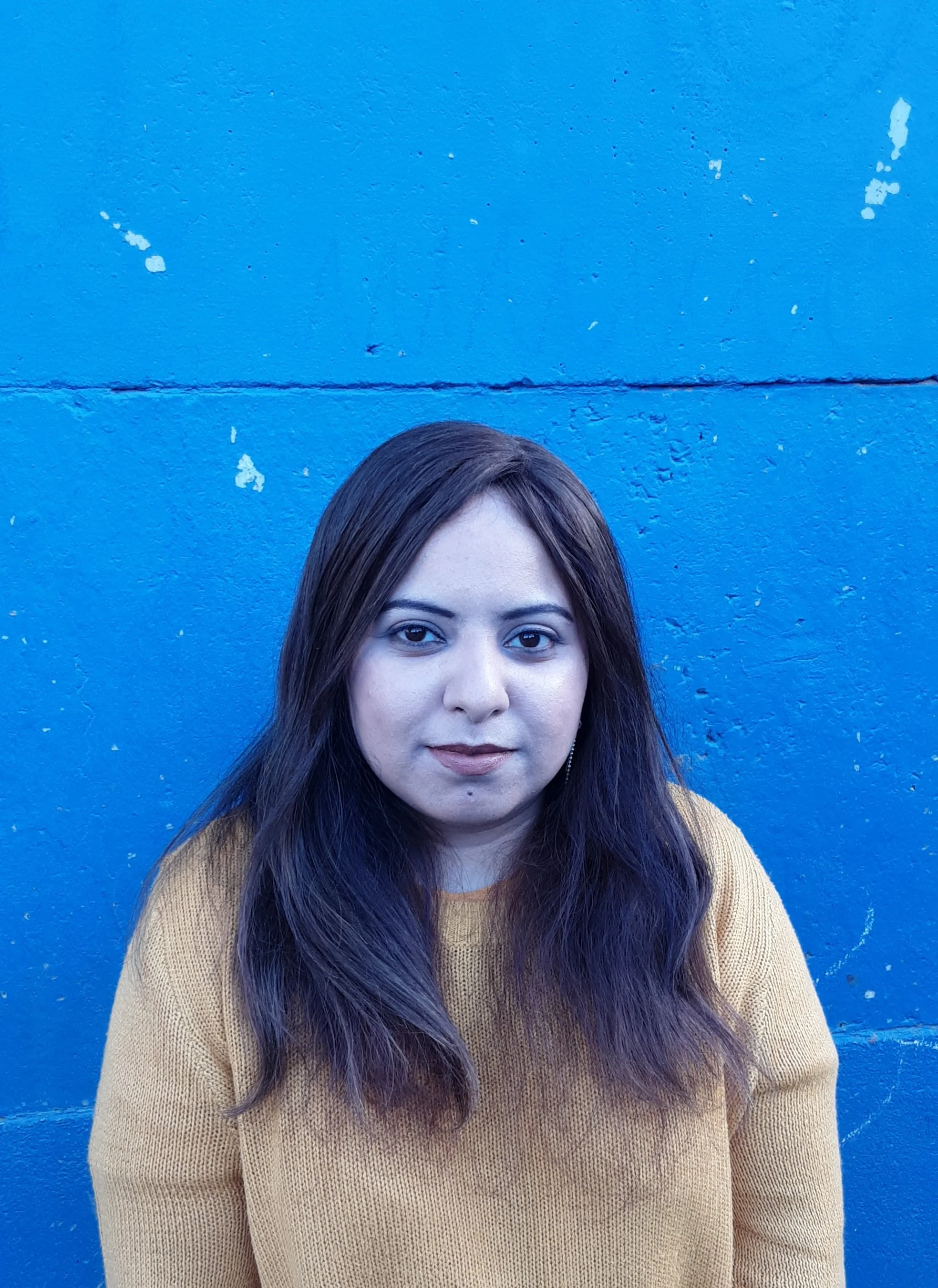 Portrait of a woman with long brown hair wearing a mustard yellow sweater standing against a blue wall.