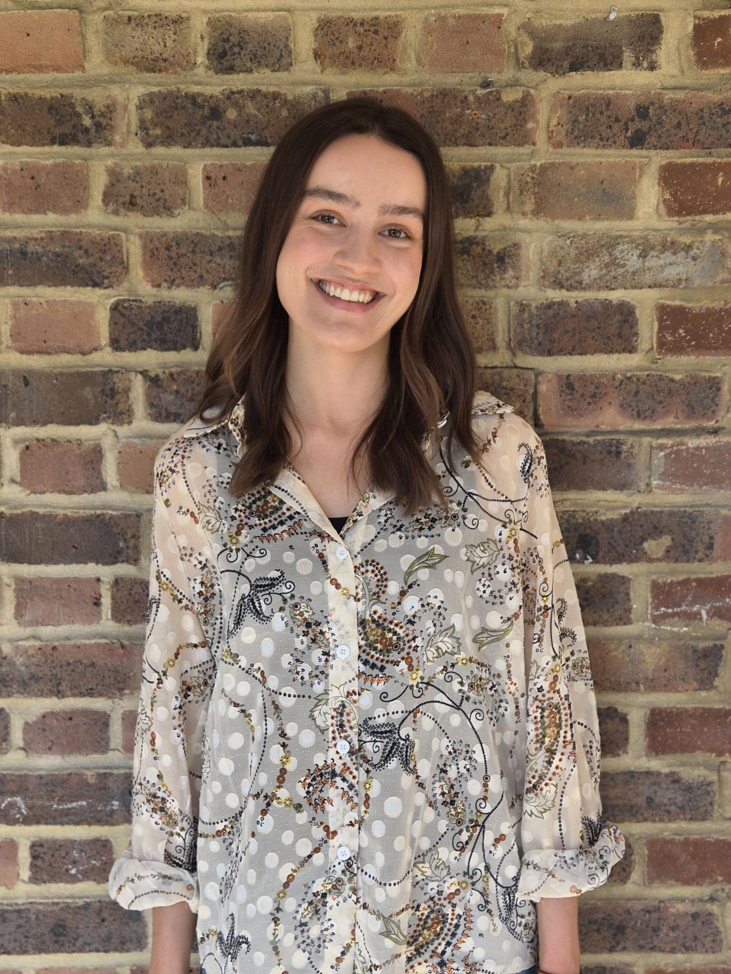 A young woman with shoulder-length brown hair, smiling, standing in front of a brick wall. She is wearing a teal cardigan and blue jeans.