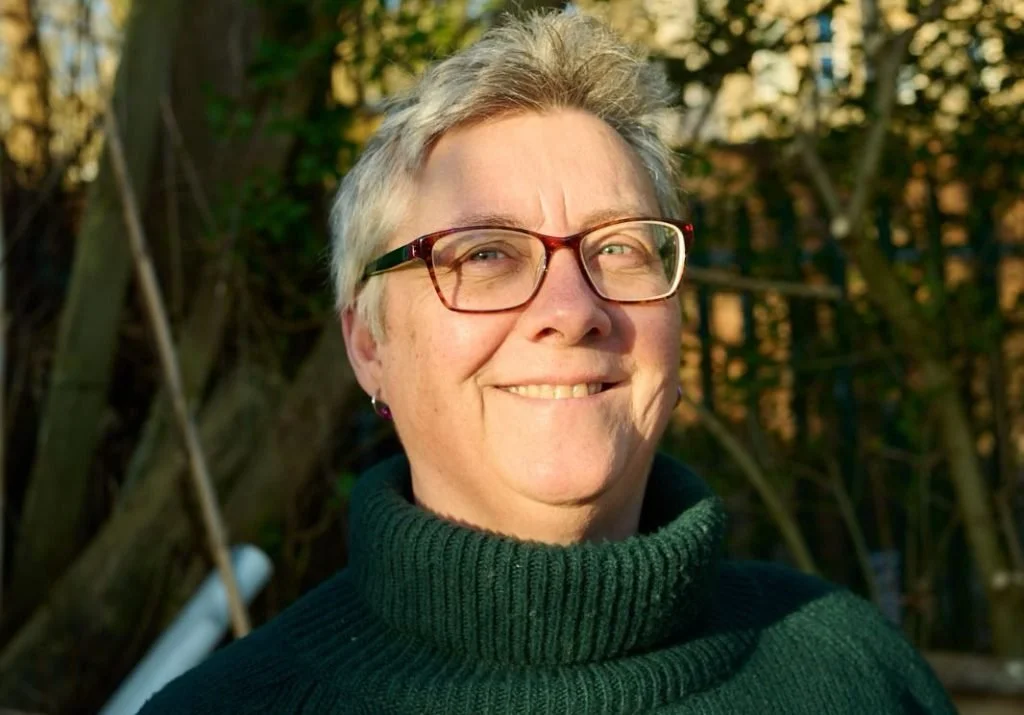A woman with short gray hair, glasses, and a dark green turtleneck sweater outdoors, smiling with a background of trees and sunlight.