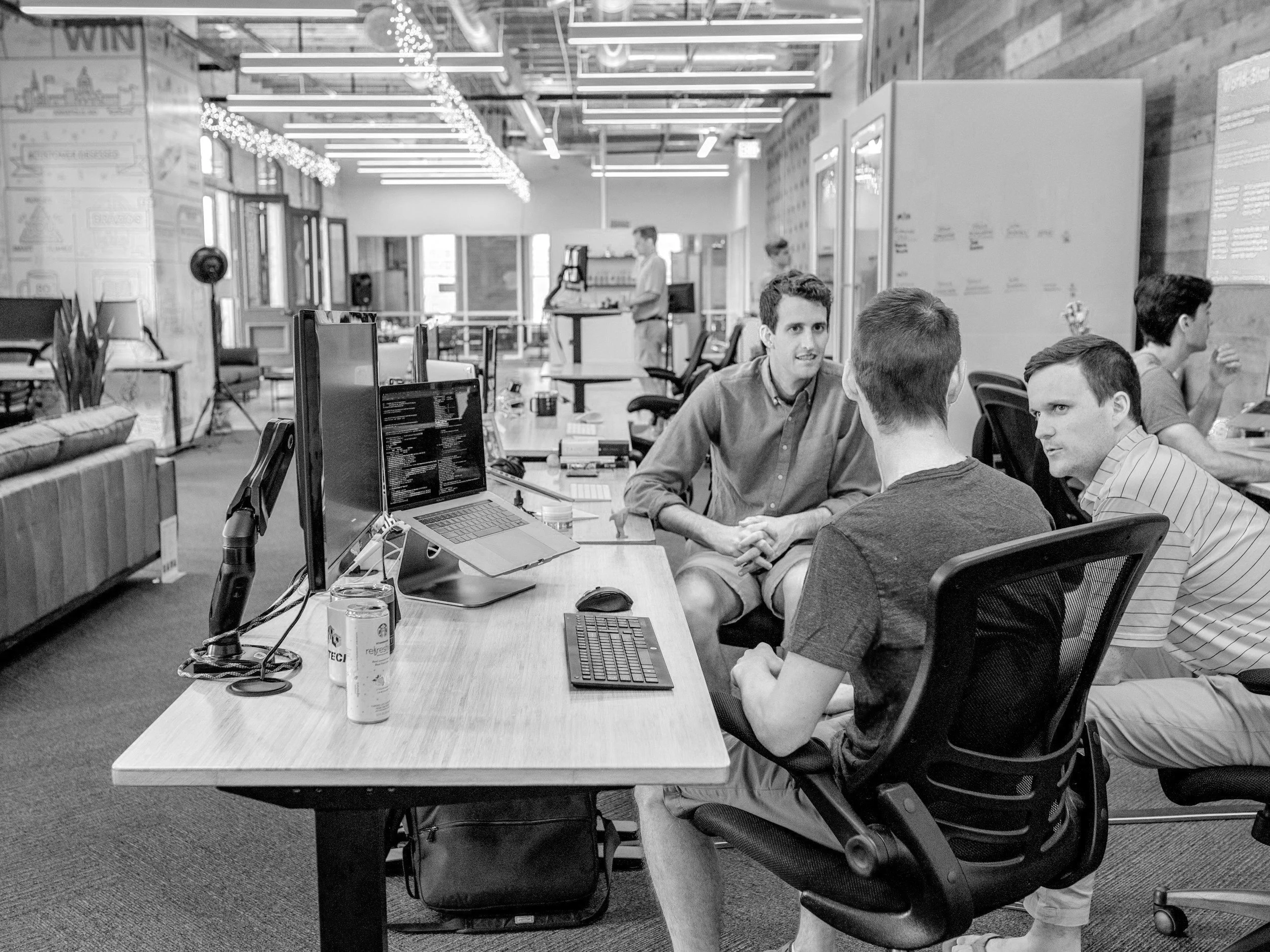Three people sitting and talking in a modern office space with computers on desks, open floor plan, and overhead lights.