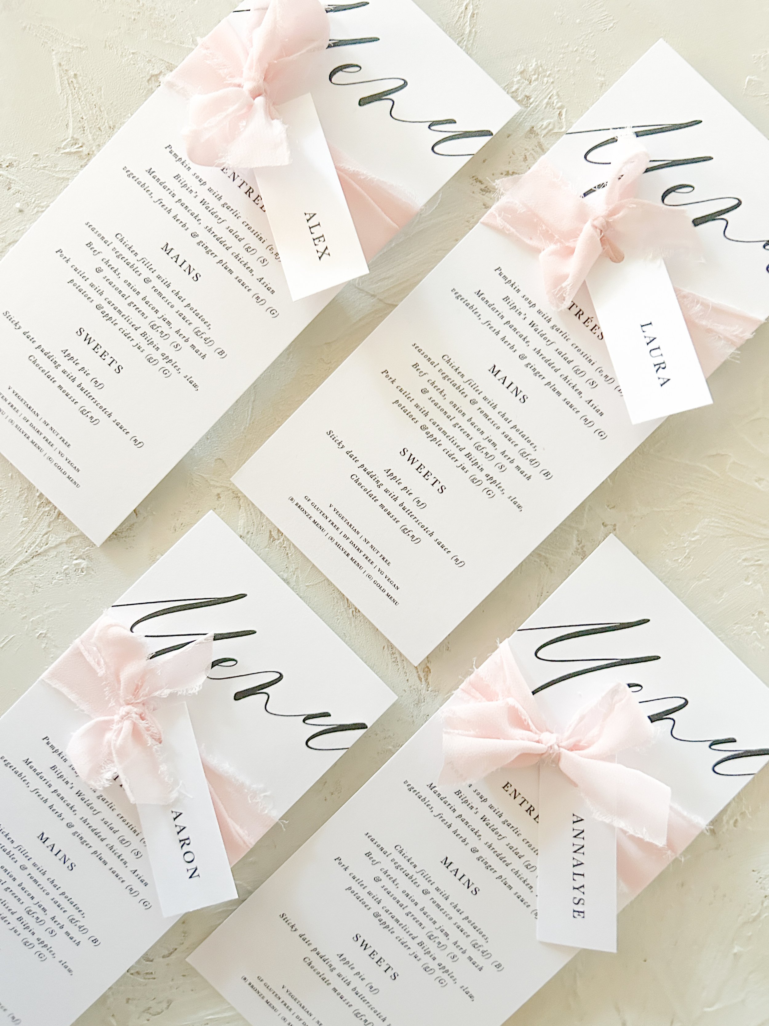 Multiple printed menus with elegant calligraphy, decorated with pink fabric bows, spread on a textured surface.