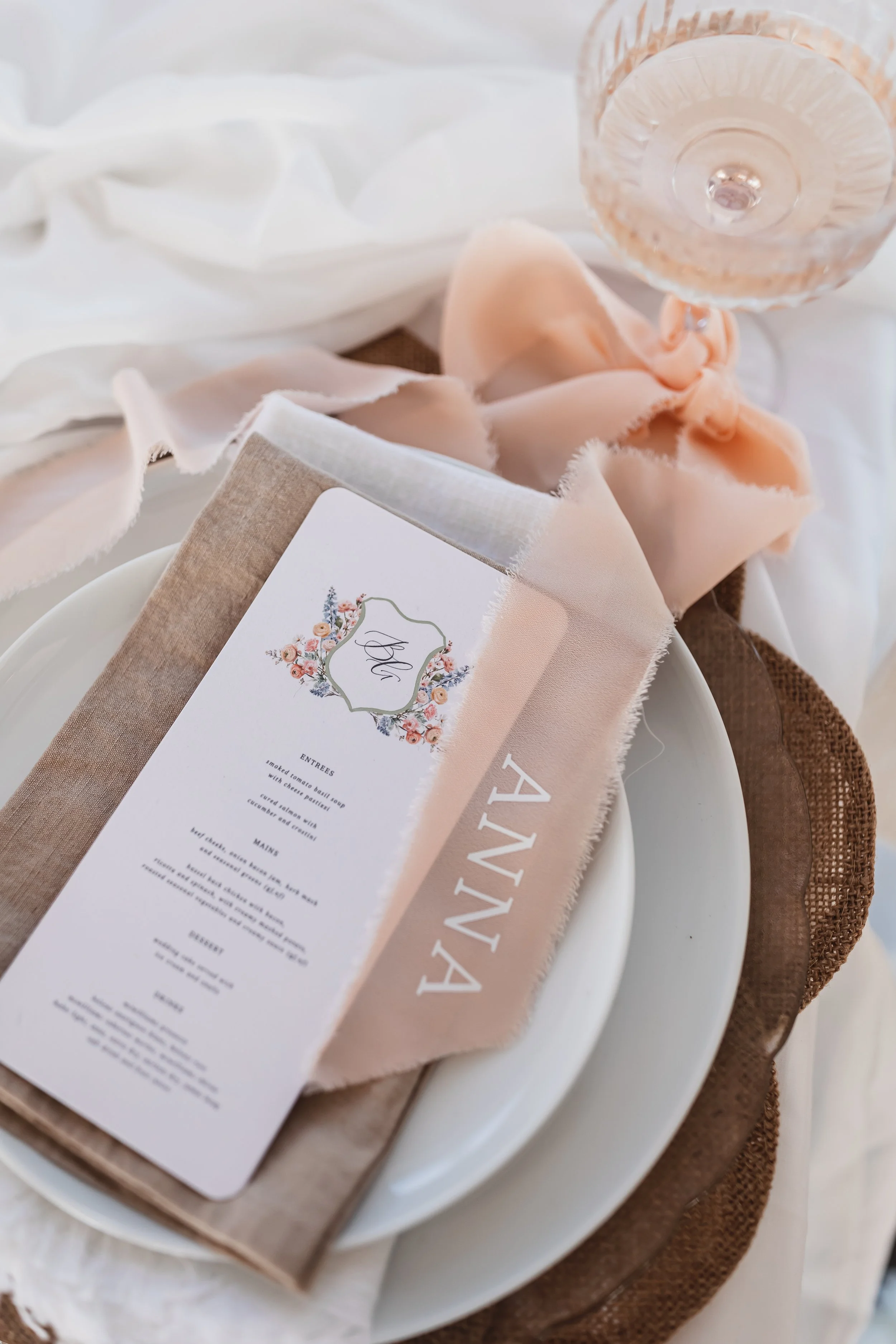A table setting with a white plate layered on top of a larger plate, a peach-colored napkin, a wine glass, and a menu with floral design in soft pastel colors.