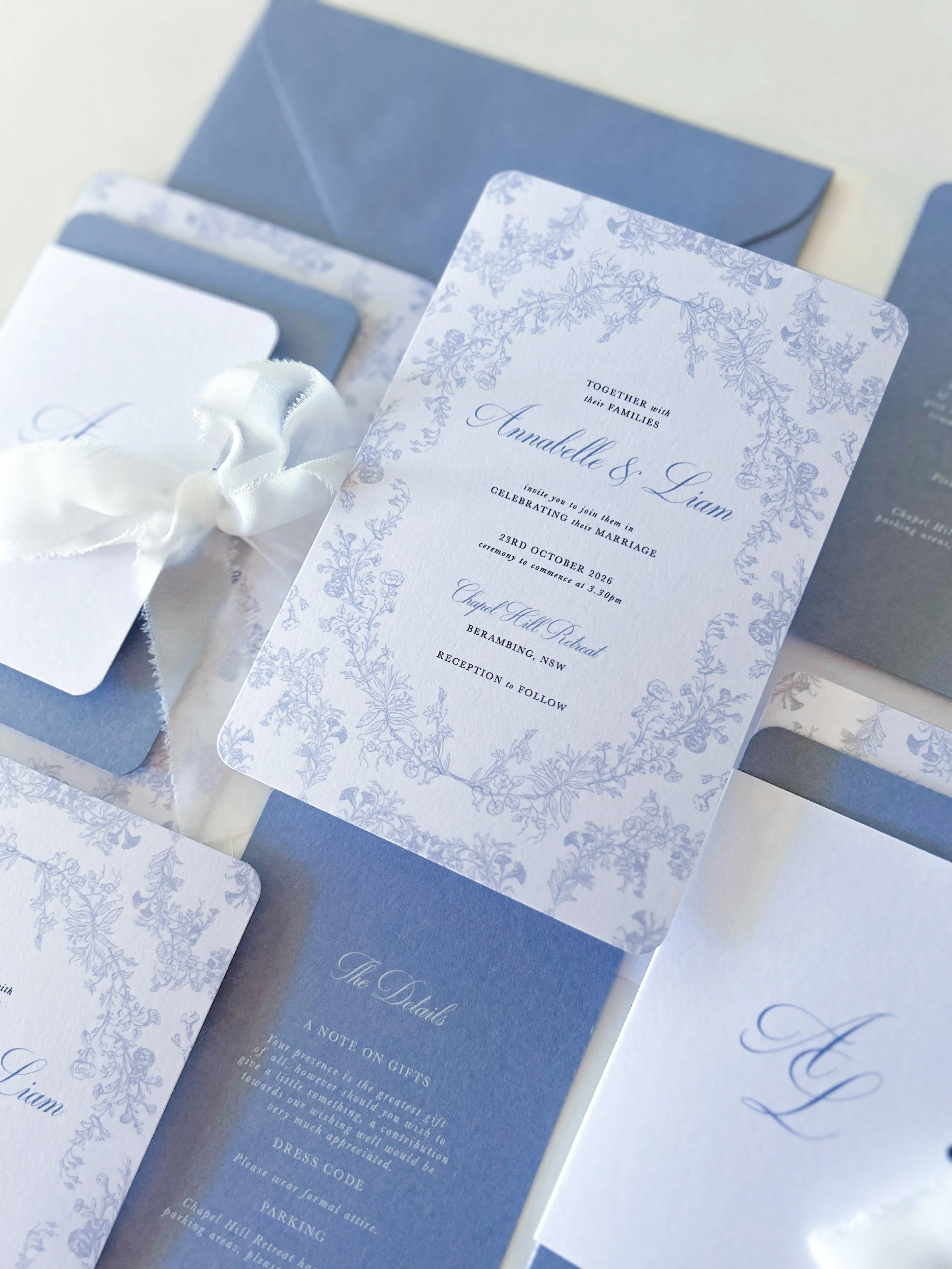 A wedding invitation with blue and white floral design, text details of the wedding, and matching envelopes, placed on a surface.