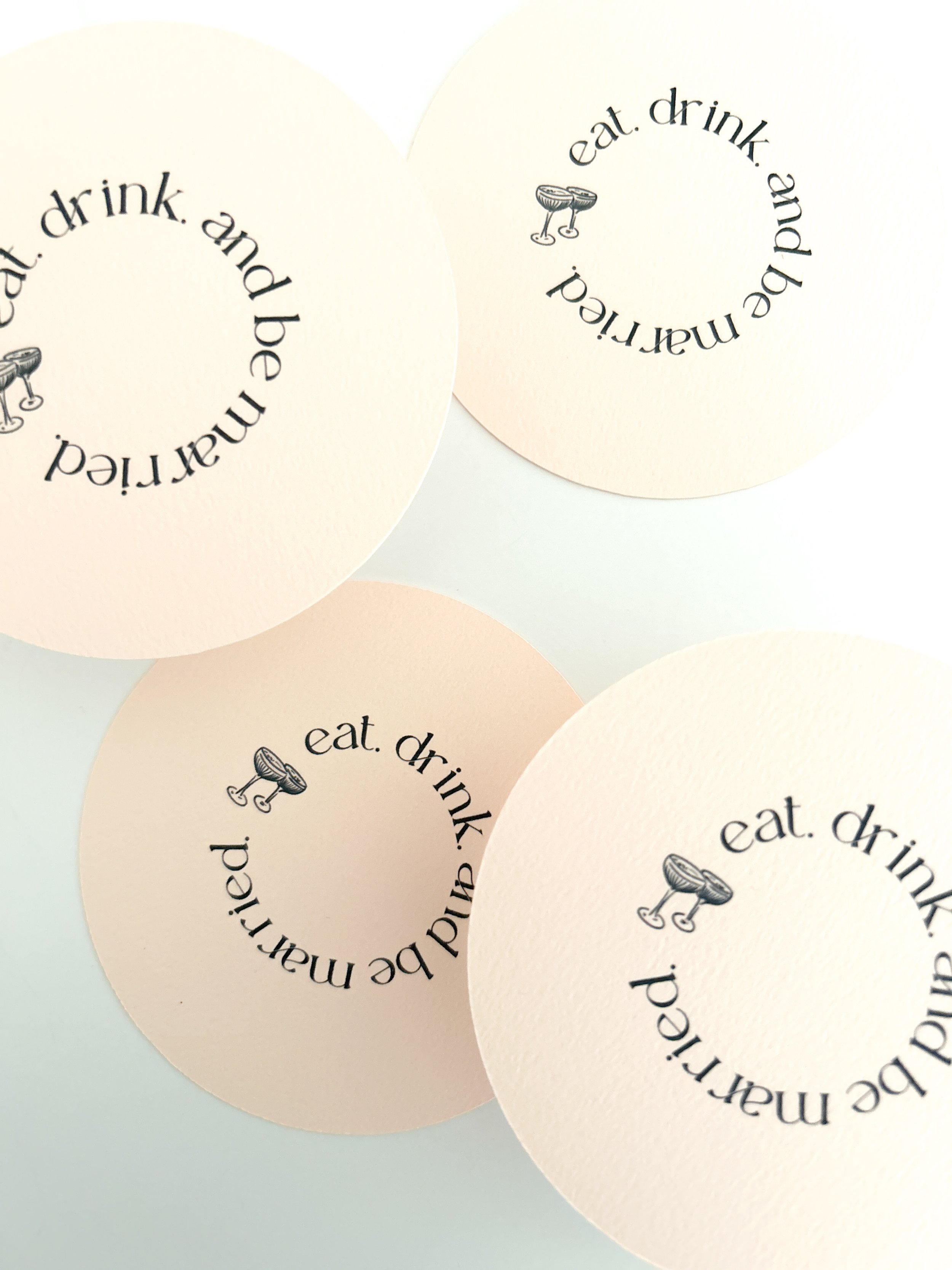 White circular paper coasters with black text and a small illustration of two drinks, reading 'eat. drink. and be merry.' arranged in a circle.