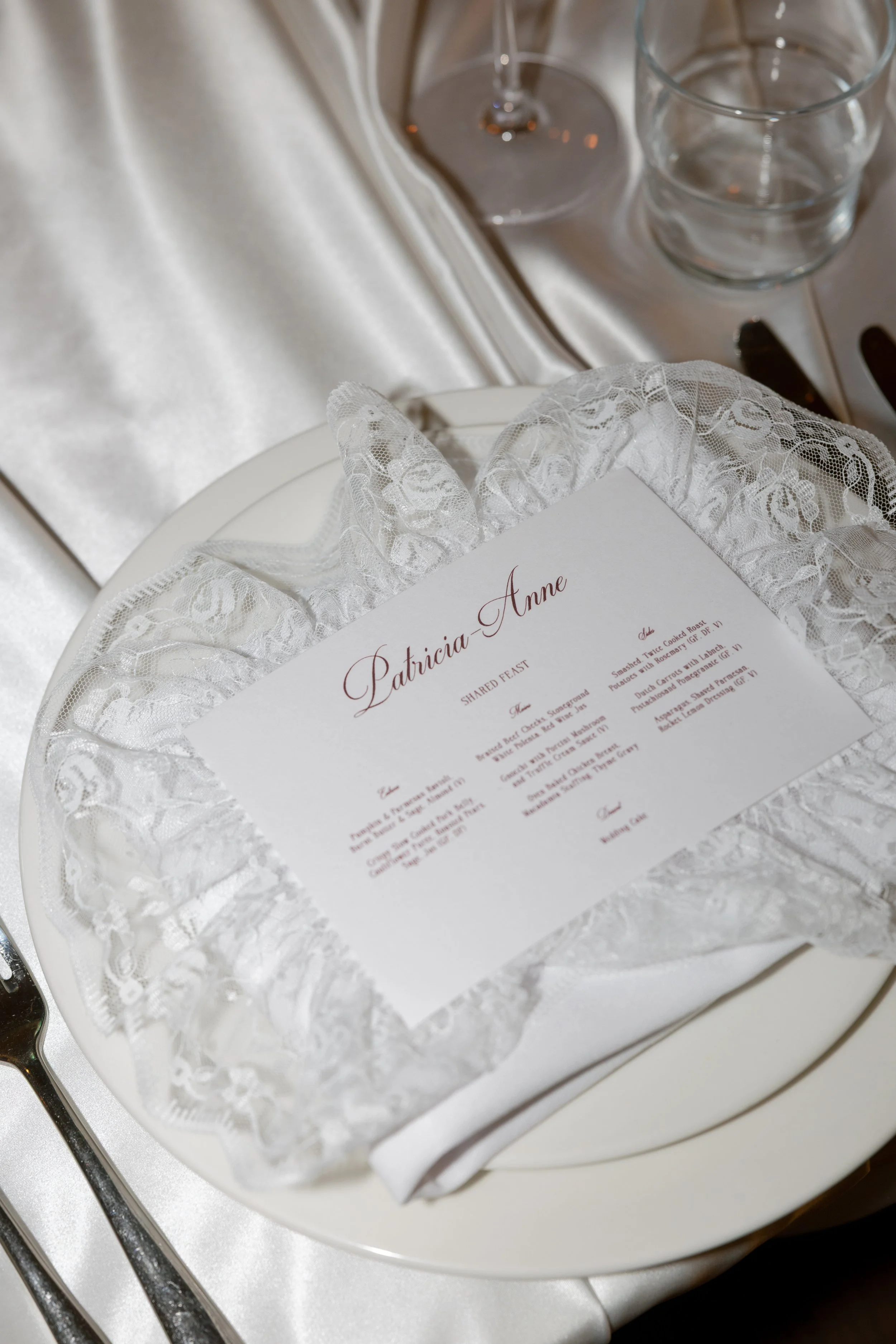 A wedding reception menu card with lace trim on a white plate, with a silver fork on the left, and a tray with empty glasses on a satin cloth in the background.