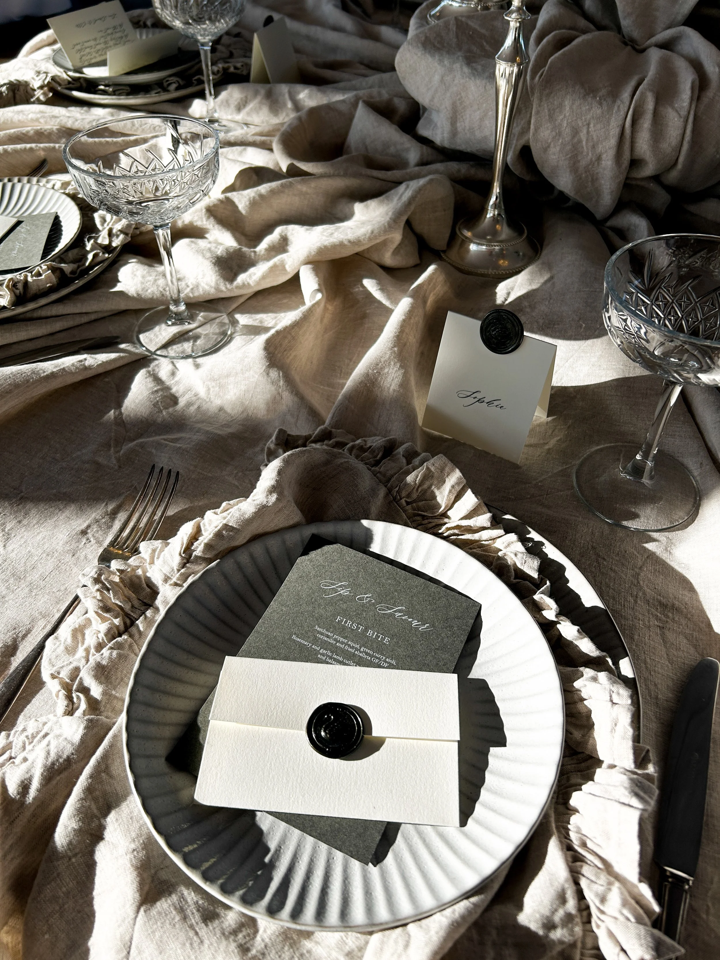 A formal table setting with a white plate on a black fluted charger plate, a menu card with a black wax seal, silverware, crystal glasses, and a name card labeled 'Sophie' on a linen tablecloth with beige and taupe fabrics.