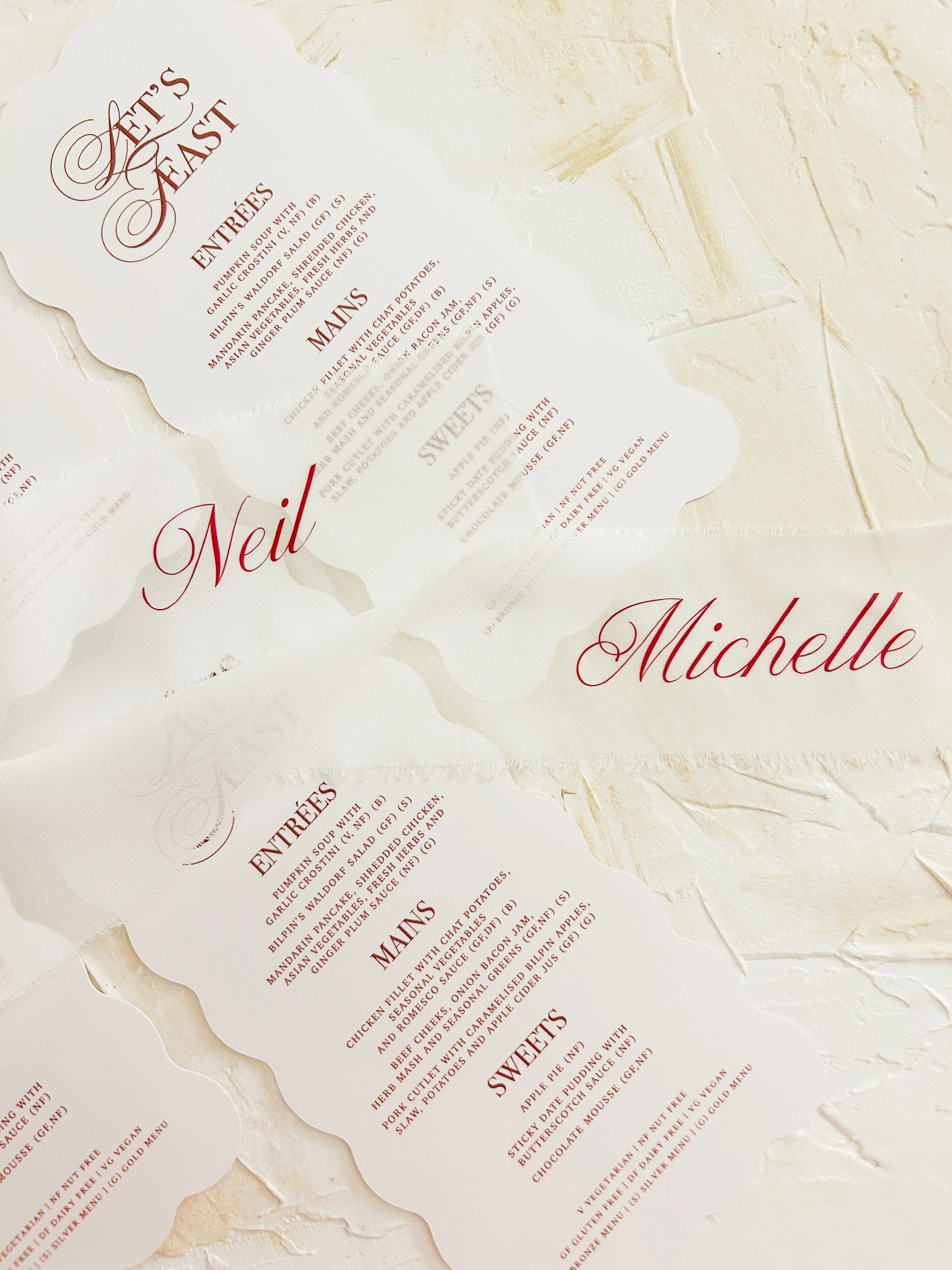 Close-up of a printed menu with a scalloped edge design, featuring sections for 'Entrees', 'Main', and 'Sweet', with the words 'Neil' and 'Michelle' in elegant red cursive font.