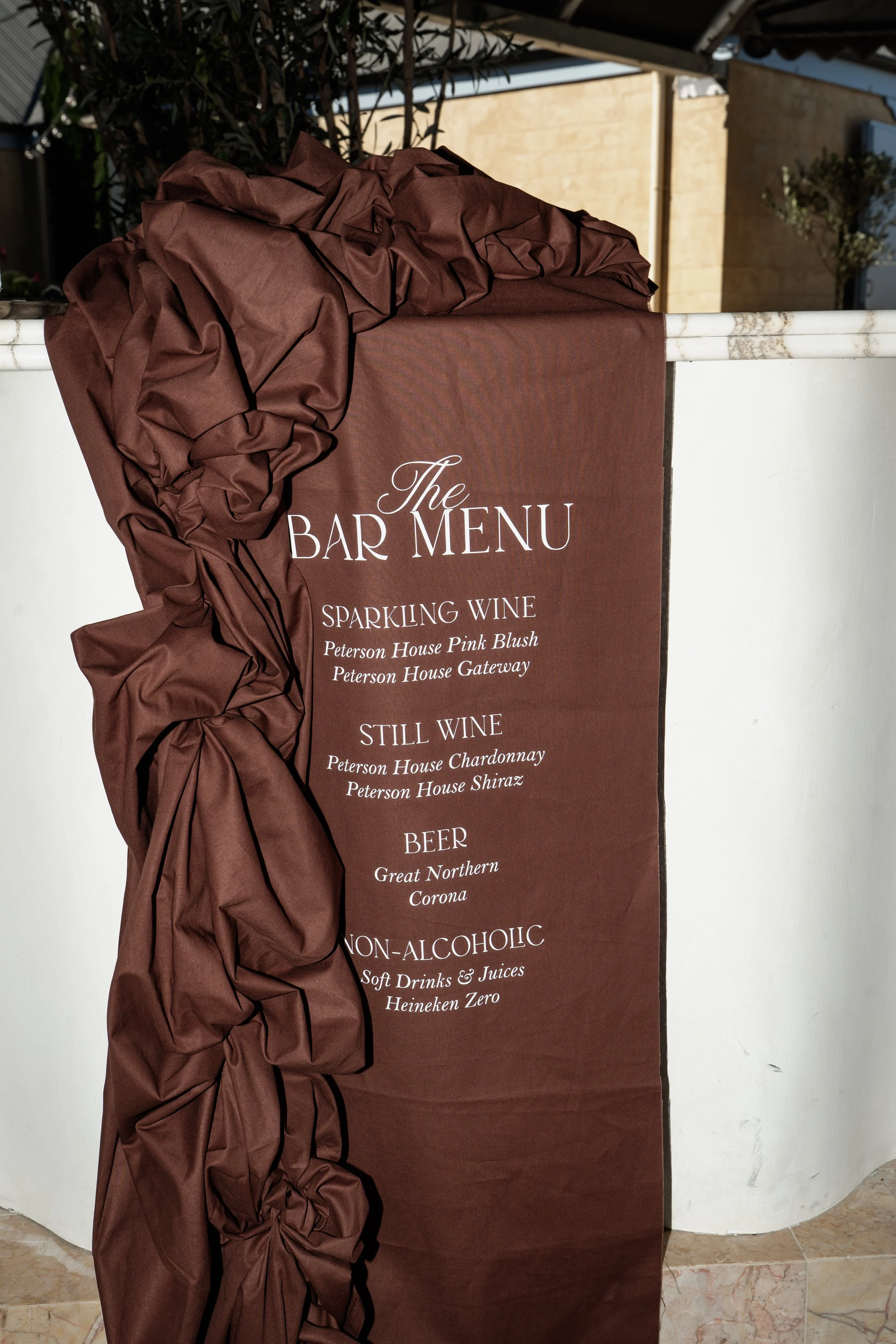 A maroon-colored banner displaying a bar menu with options for sparkling wine, still wine, beer, and non-alcoholic drinks. The banner is attached to a white surface and decorated with crumpled brown fabric.