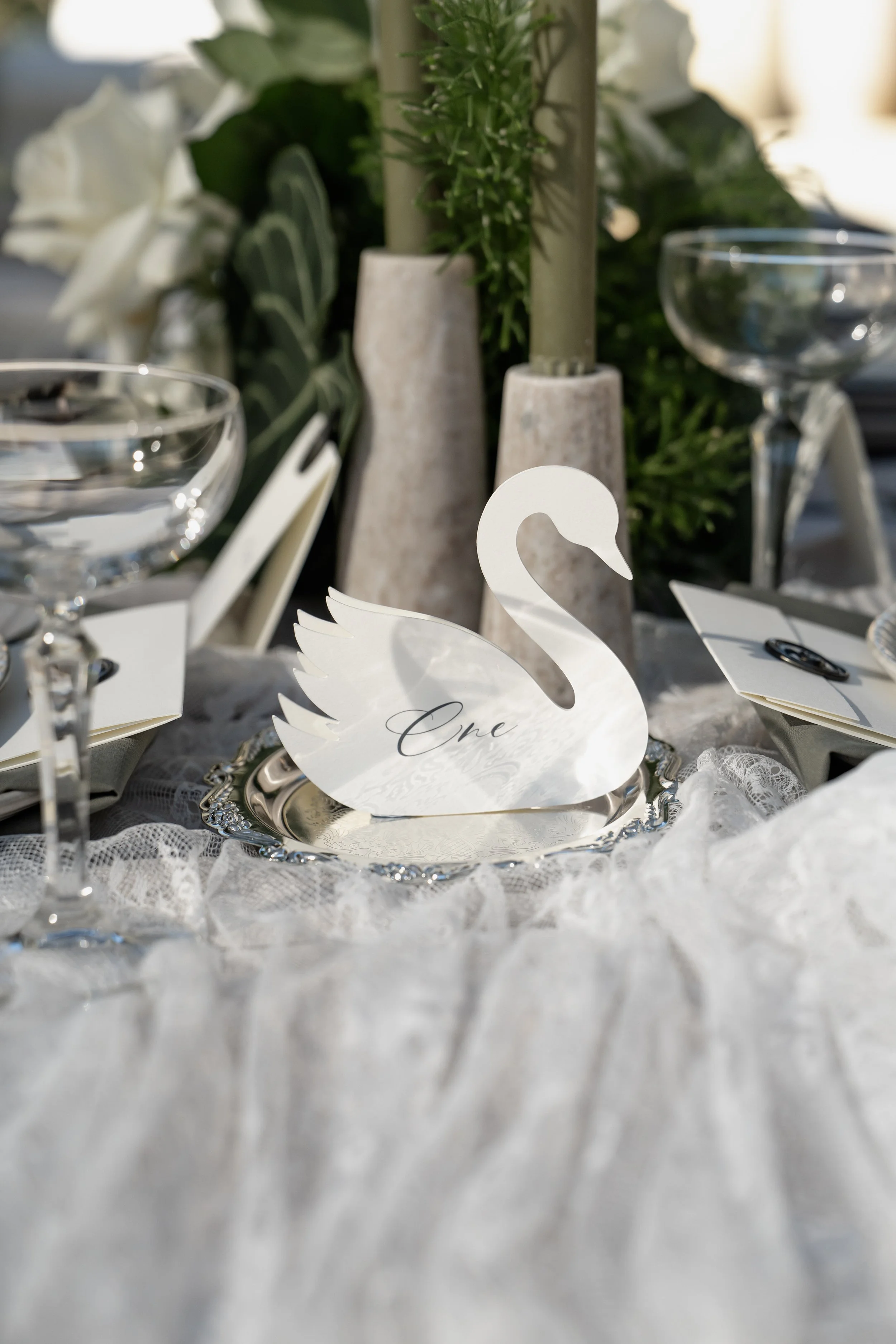 Elegant table setting with a white swan-shaped place card that says "One," surrounded by crystal glasses, white lace tablecloth, and decorative greenery in the background.
