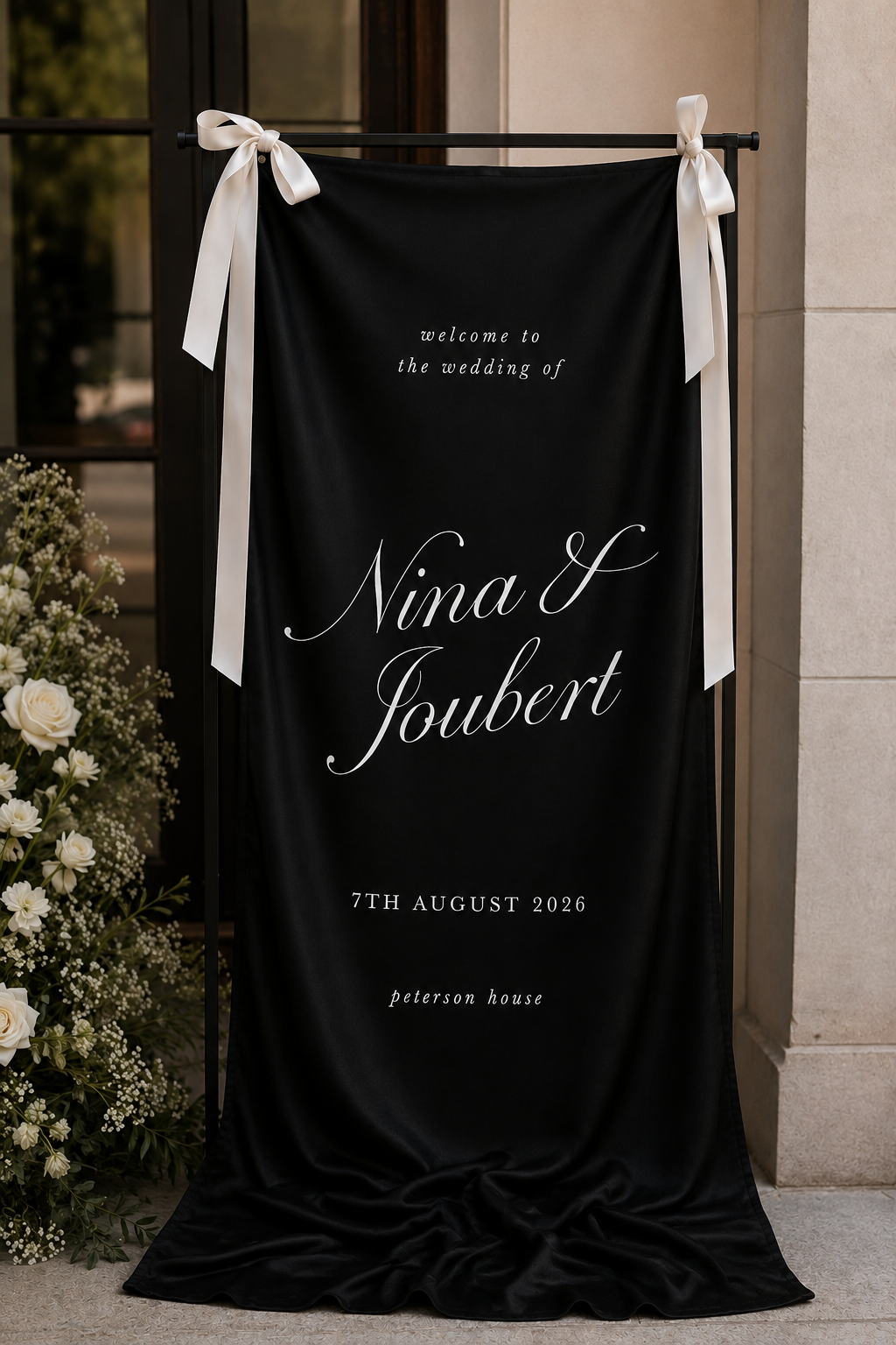 A black fabric wedding sign with white cursive text welcoming guests to the wedding of Nina V Joubert on August 7, 2026, at Peterson House. The sign is decorated with white ribbons on a stand, with white flowers nearby.