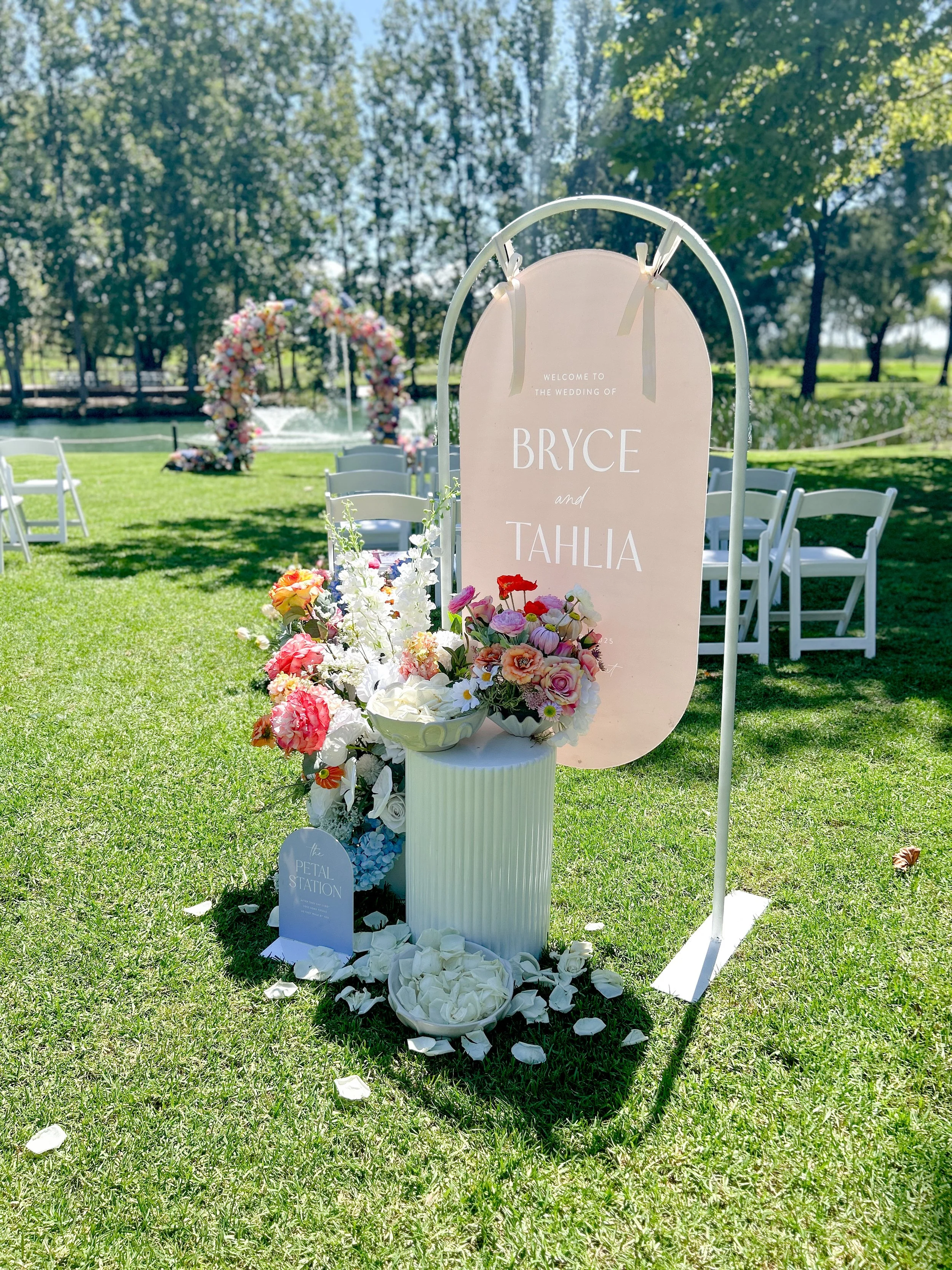 A wedding sign with pink and white flowers in front of a grassy outdoor setting with trees, chairs, and a floral arch in the background.