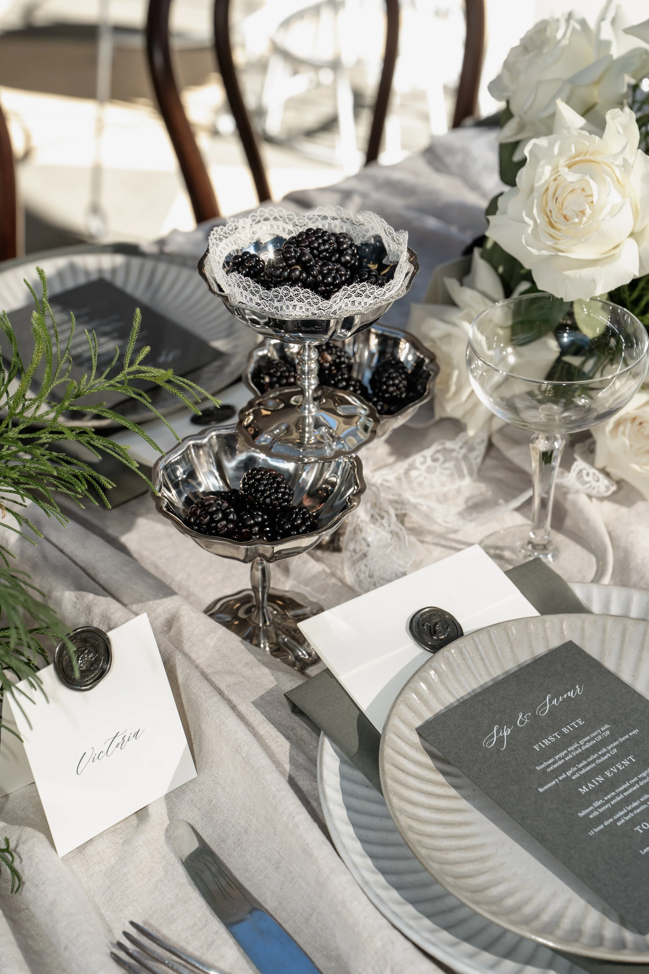 A table setting for a formal event with blackberries in silver bowls, white roses as a centerpiece, and menus with elegant calligraphy, all arranged on a white tablecloth.