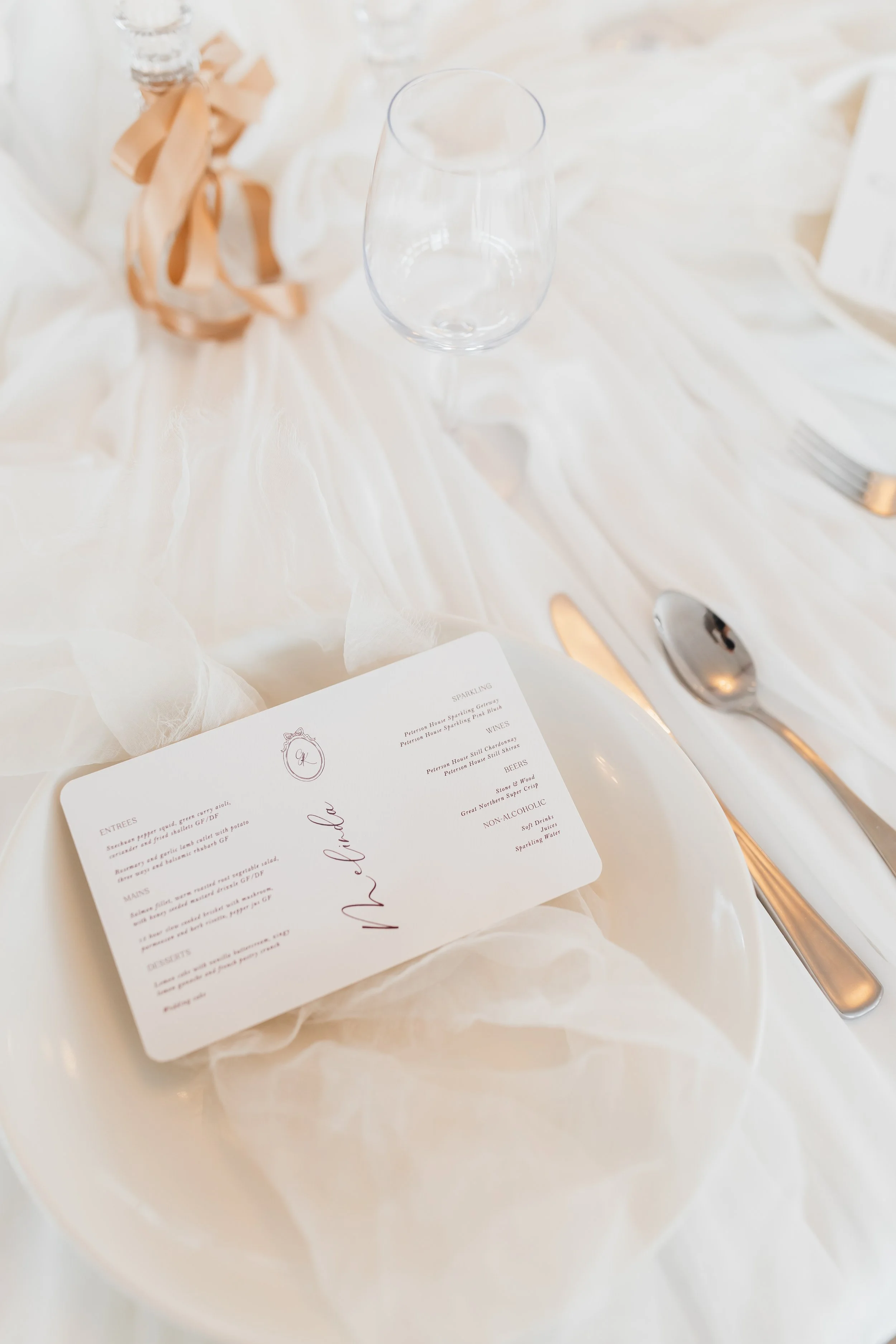 A clear wine glass on a white tablecloth next to a beige flatware and a plate with a menu card. The menu features a list of entrees, mains, desserts, and beverages in elegant script, with a decorative monogram at the top.