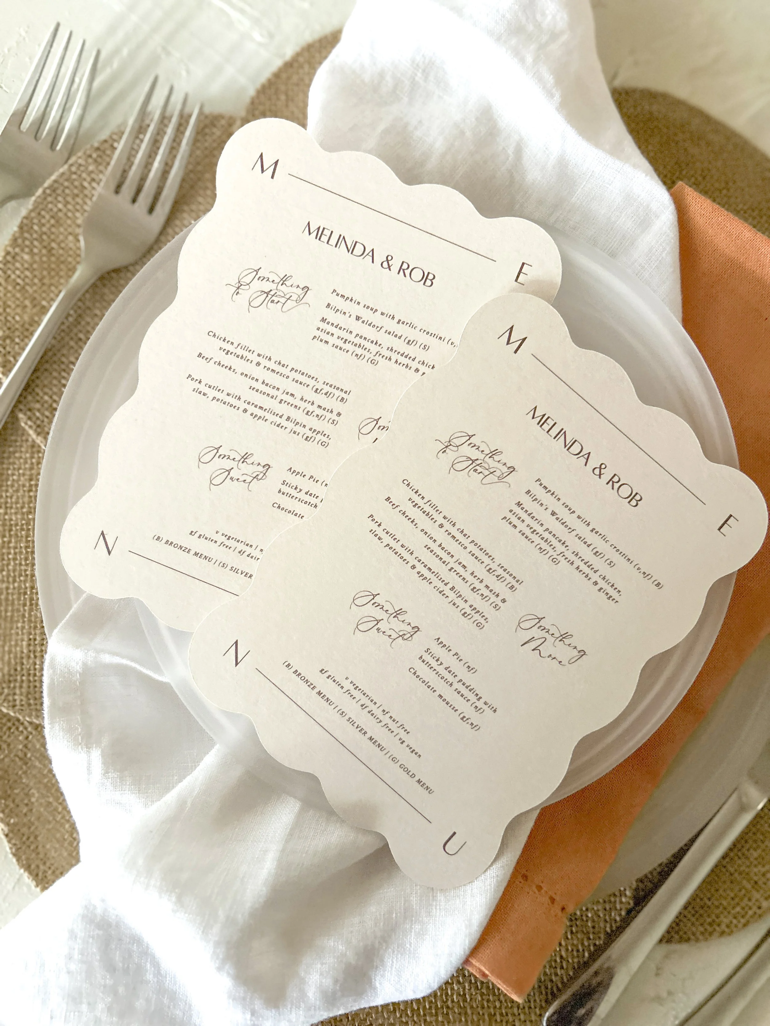 A table setting featuring a white plate with a paper menu, silver fork, brown napkin, and an orange napkin on a white tablecloth.