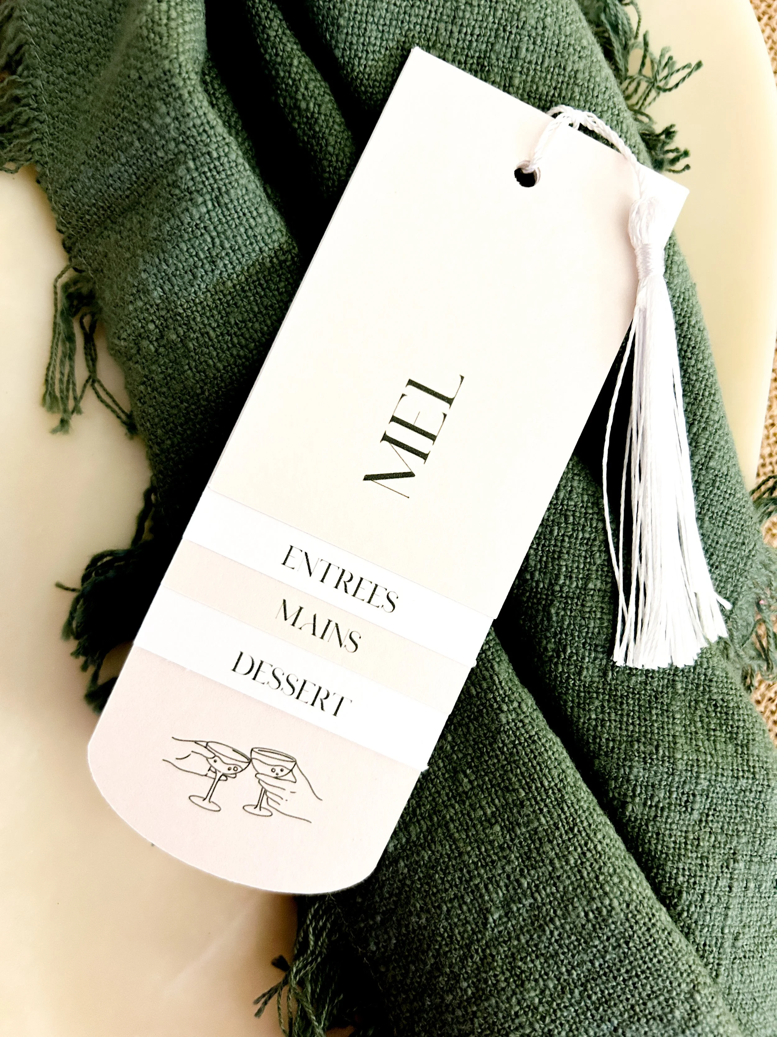 Close-up of a white menu card with black text, resting on a green textured scarf, with a cartoon illustration of two clinking glasses at the bottom of the menu.