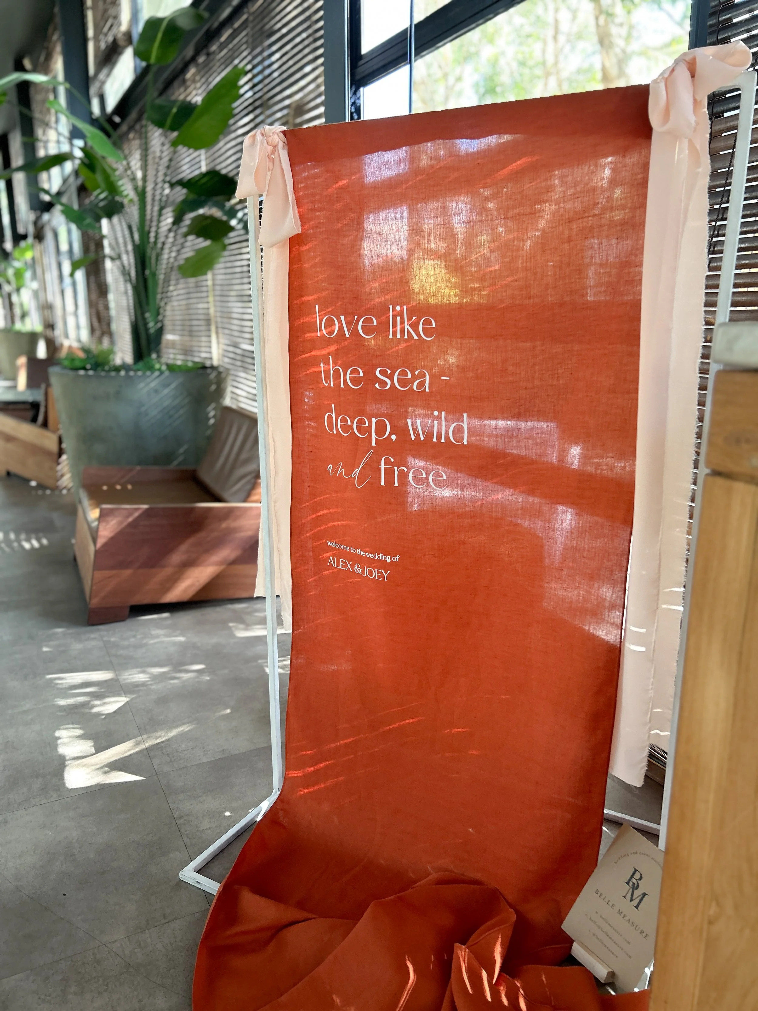 A red fabric banner with white text that reads 'love like the sea - deep, wild and free' standing next to a mirror and a potted plant inside a room with wooden and metal elements.