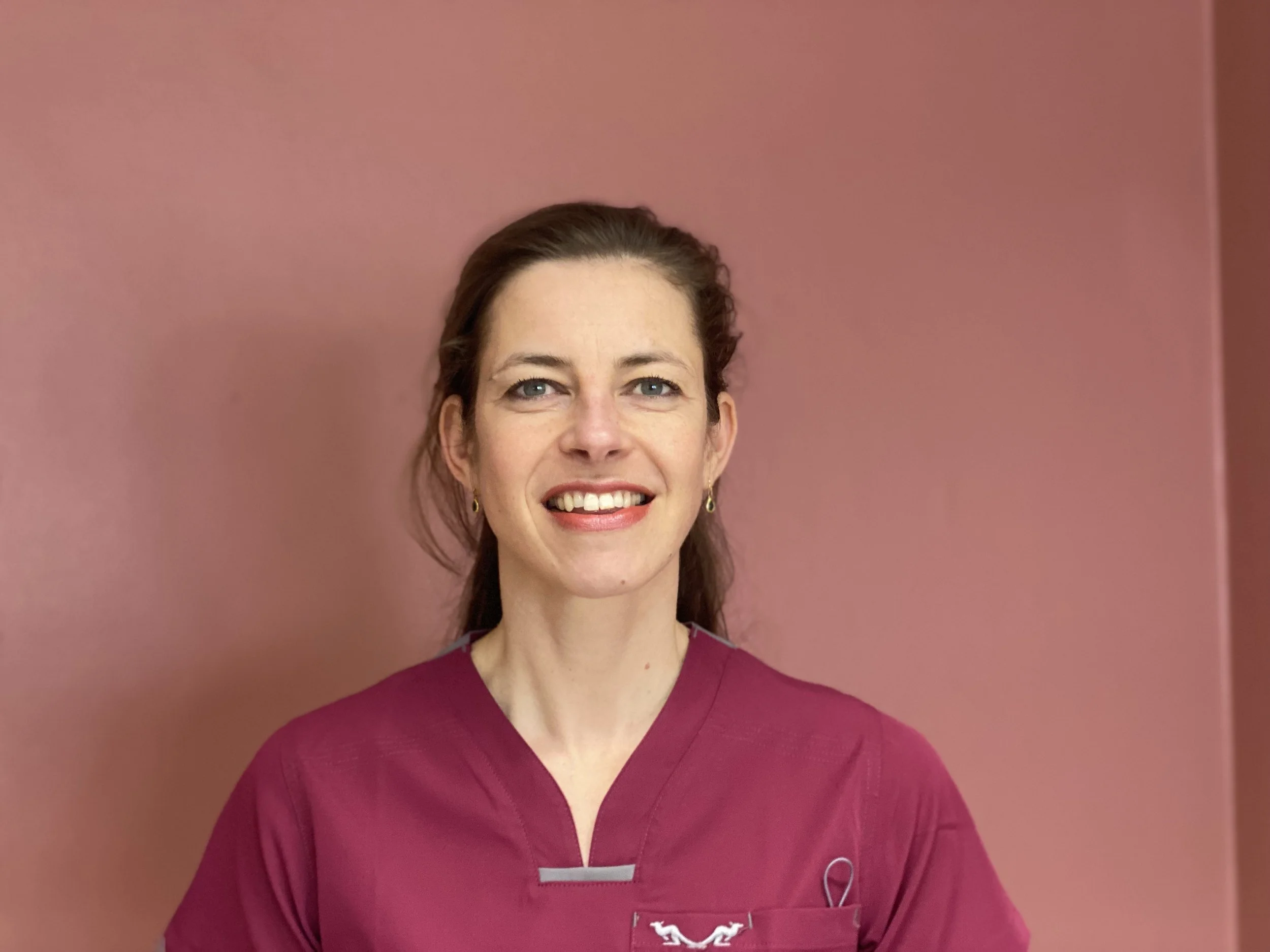 Portrait of a smiling woman with brown hair wearing a maroon medical uniform, standing against a pink wall.