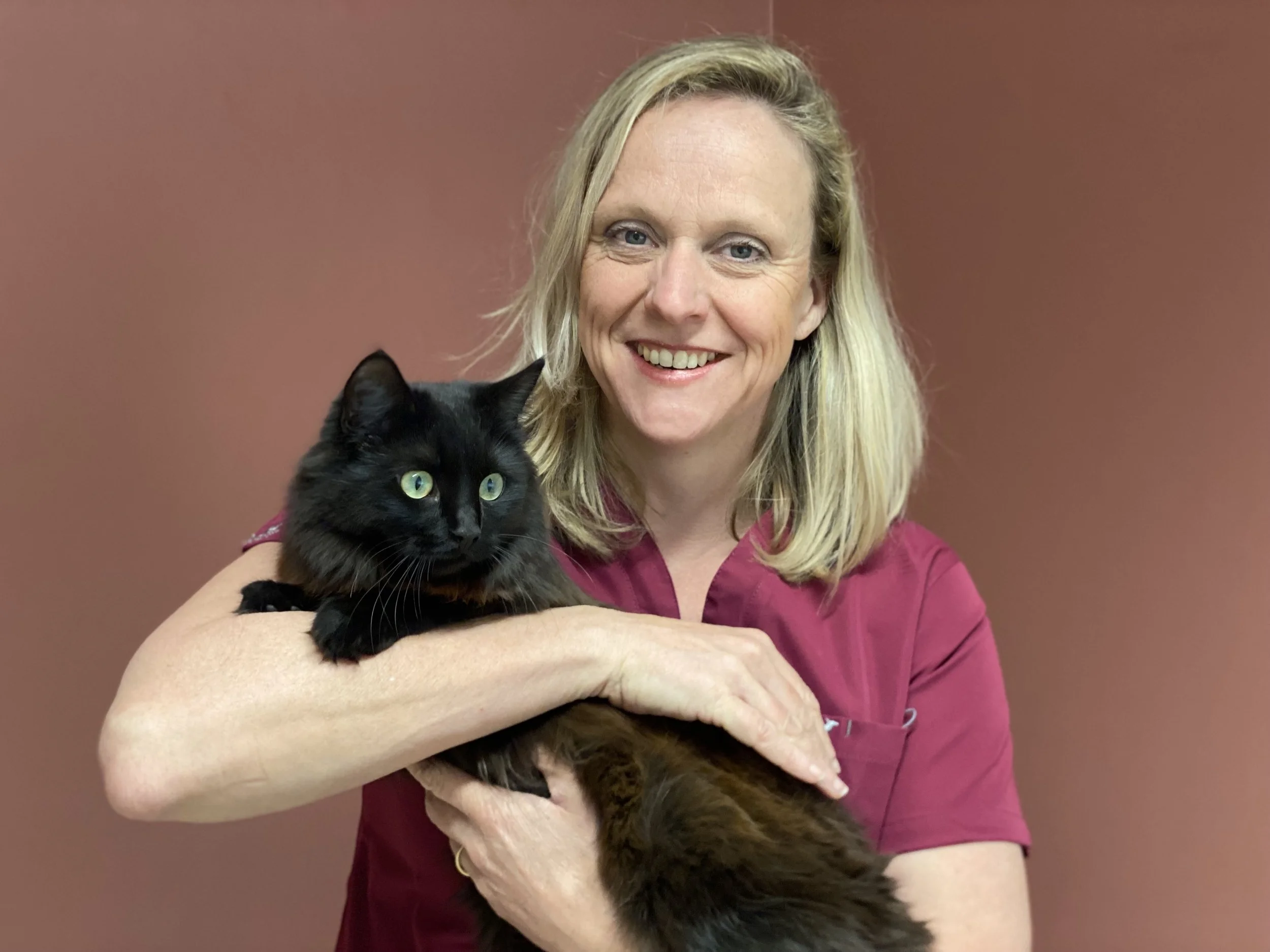 A woman in a pink shirt holding a black cat with green eyes against a plain brown background.