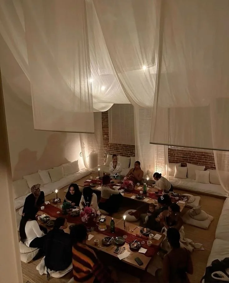 People gathered around a long dining table in a cozy, warmly lit room with exposed brick walls and draped white fabric hanging from the ceiling.