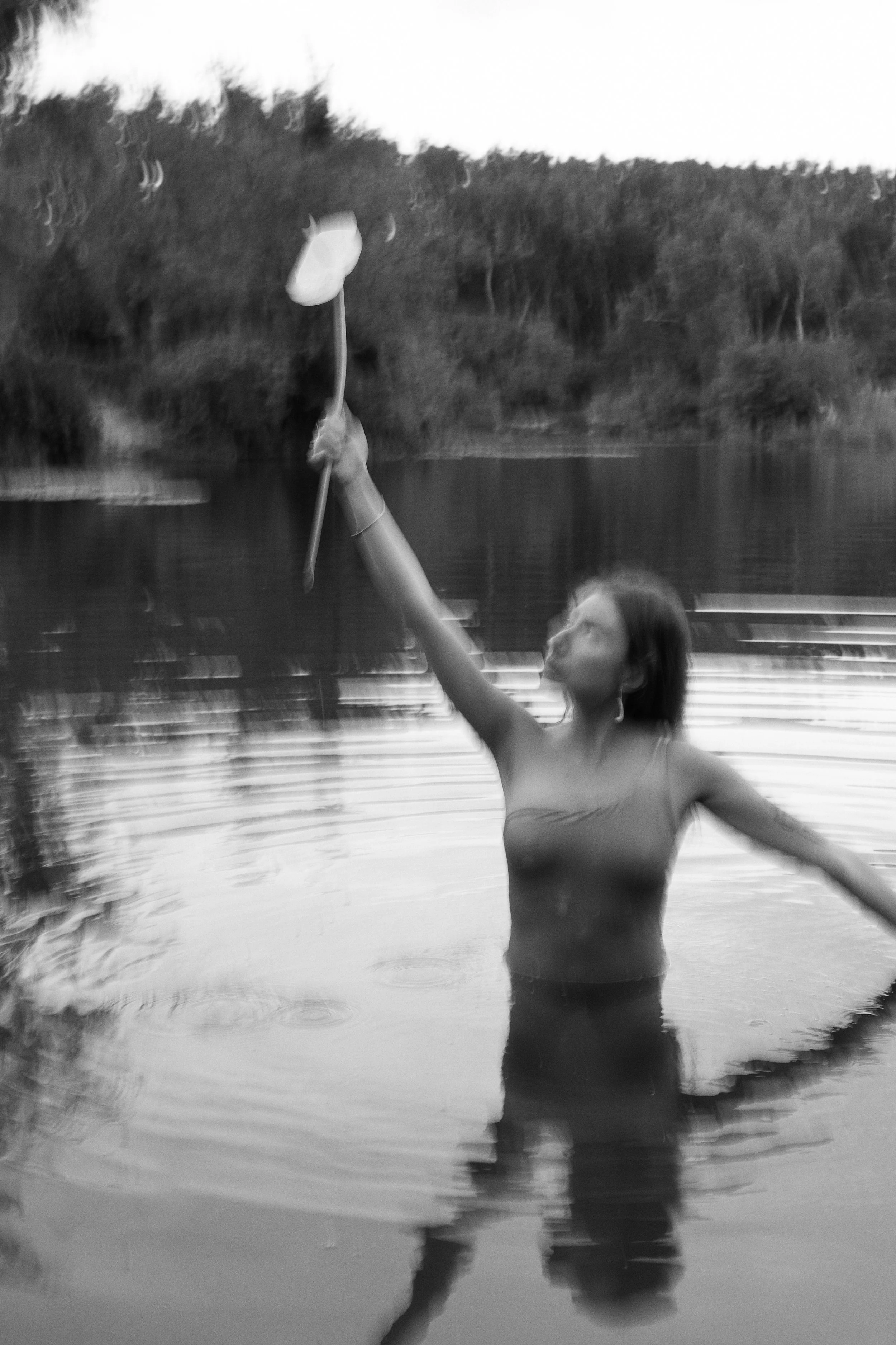 A woman in water holding a paddle above her head, with trees and a dock in the background