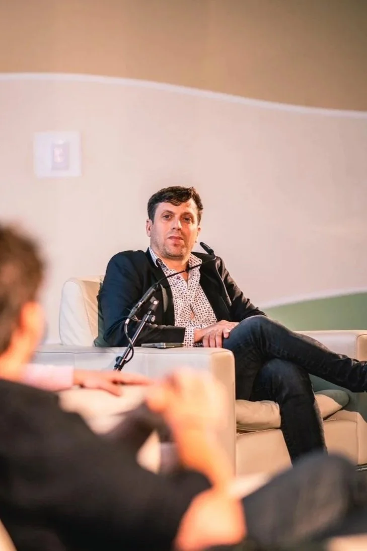 A man is sitting on a beige sofa with his legs crossed, wearing a black jacket and patterned shirt, participating in a discussion or interview, with a blurred person in the foreground.