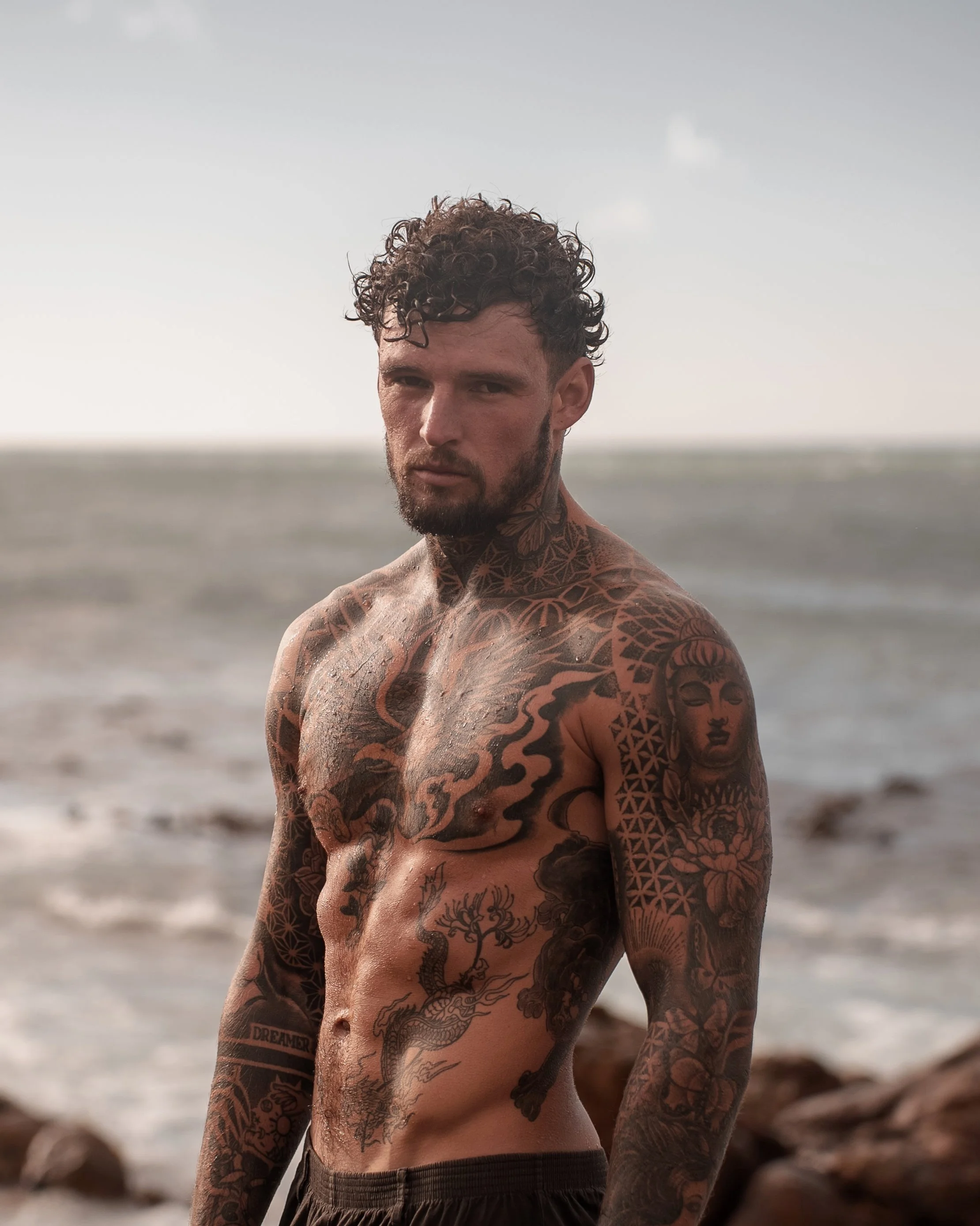 A shirtless man with tattoos on his chest, arms, and neck standing on a beach with the ocean in the background