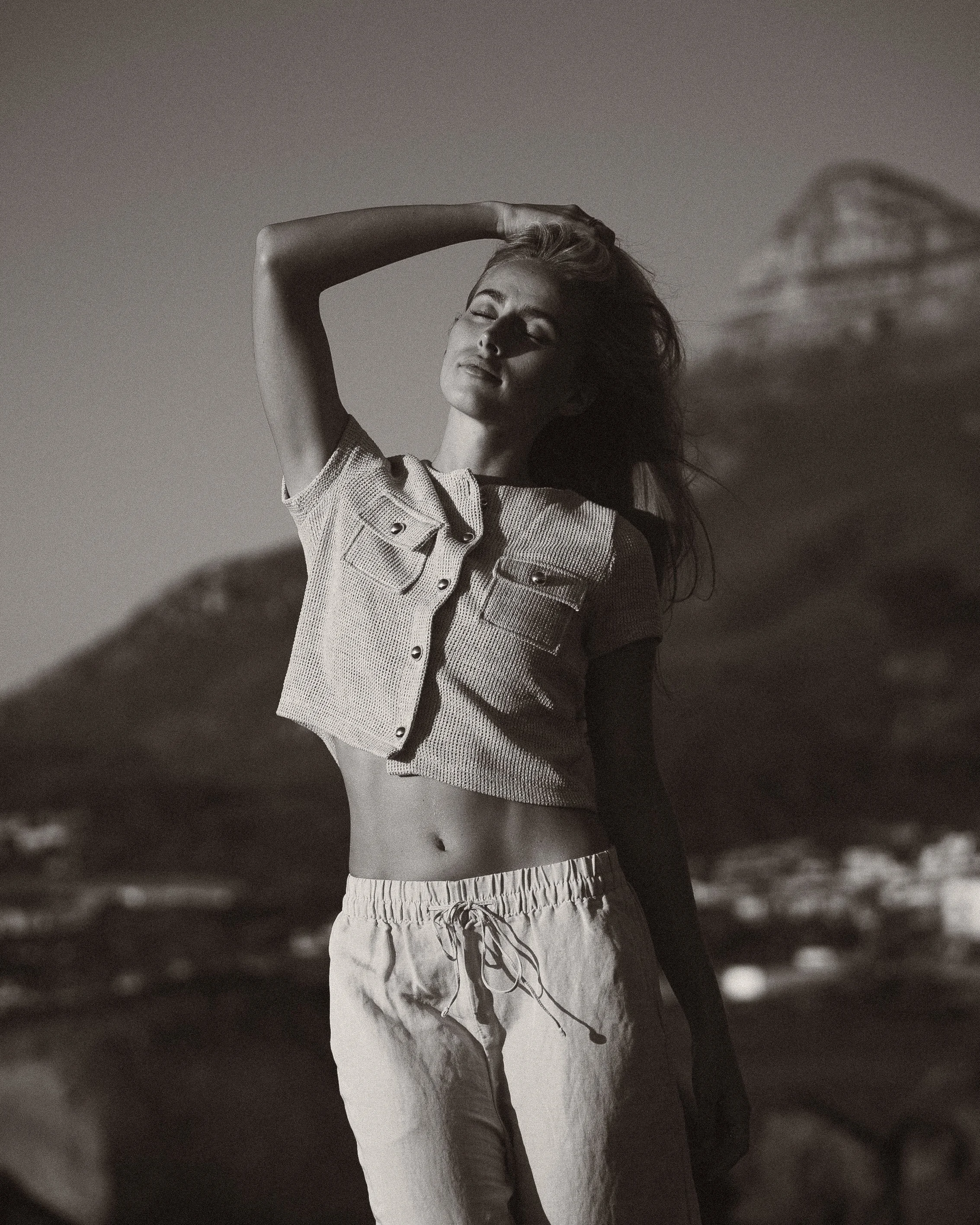 A woman posing outdoors with her eyes closed, wearing a cropped short-sleeve button-up shirt and drawstring pants, with a mountain and sky in the background.