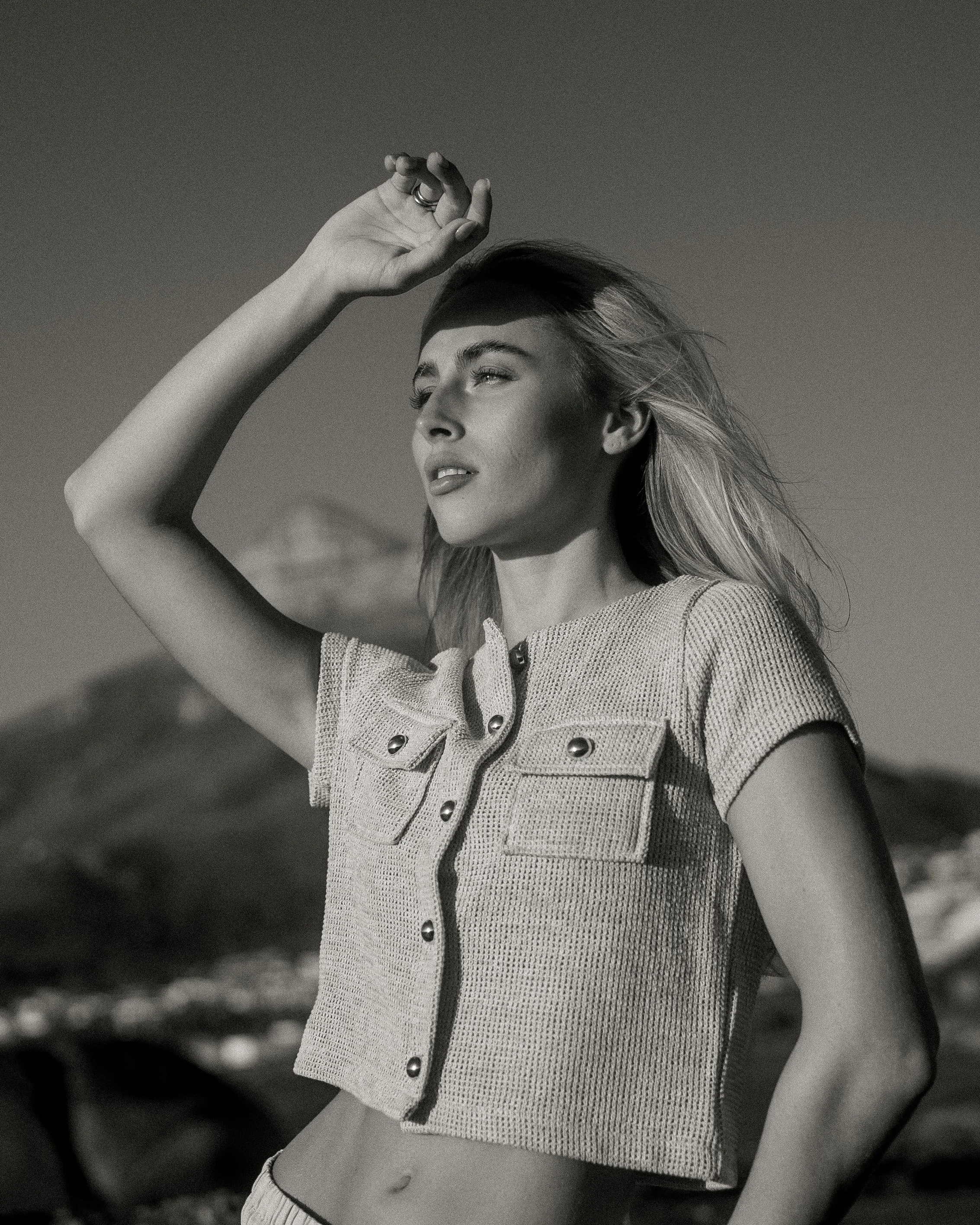 A woman with long hair, wearing a short-sleeved textured top, stands outdoors with her arm raised, shielding her eyes from the sun, in a black and white photograph.