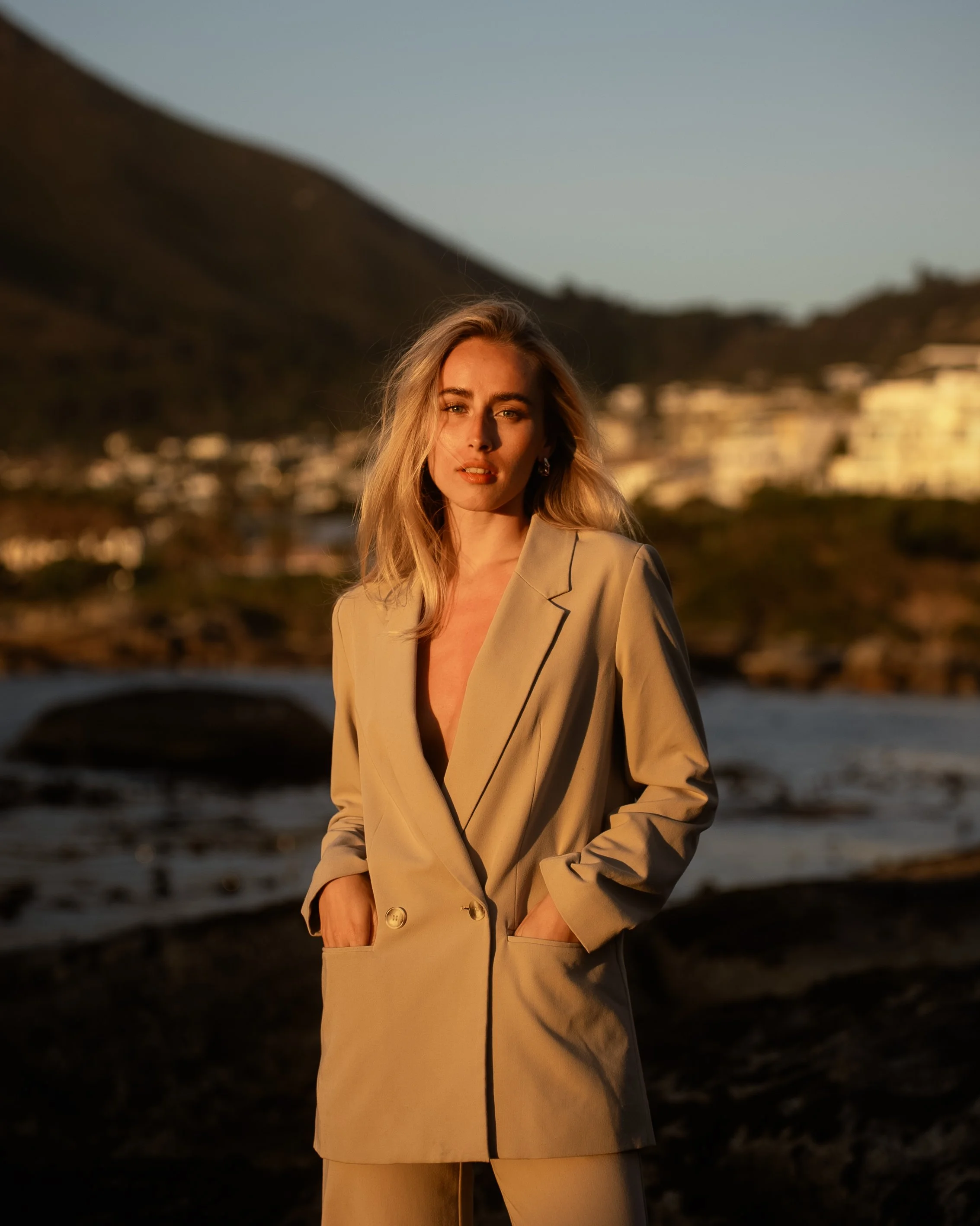 A woman in a beige blazer and pants standing outdoors during sunset with a mountain and water in the background.