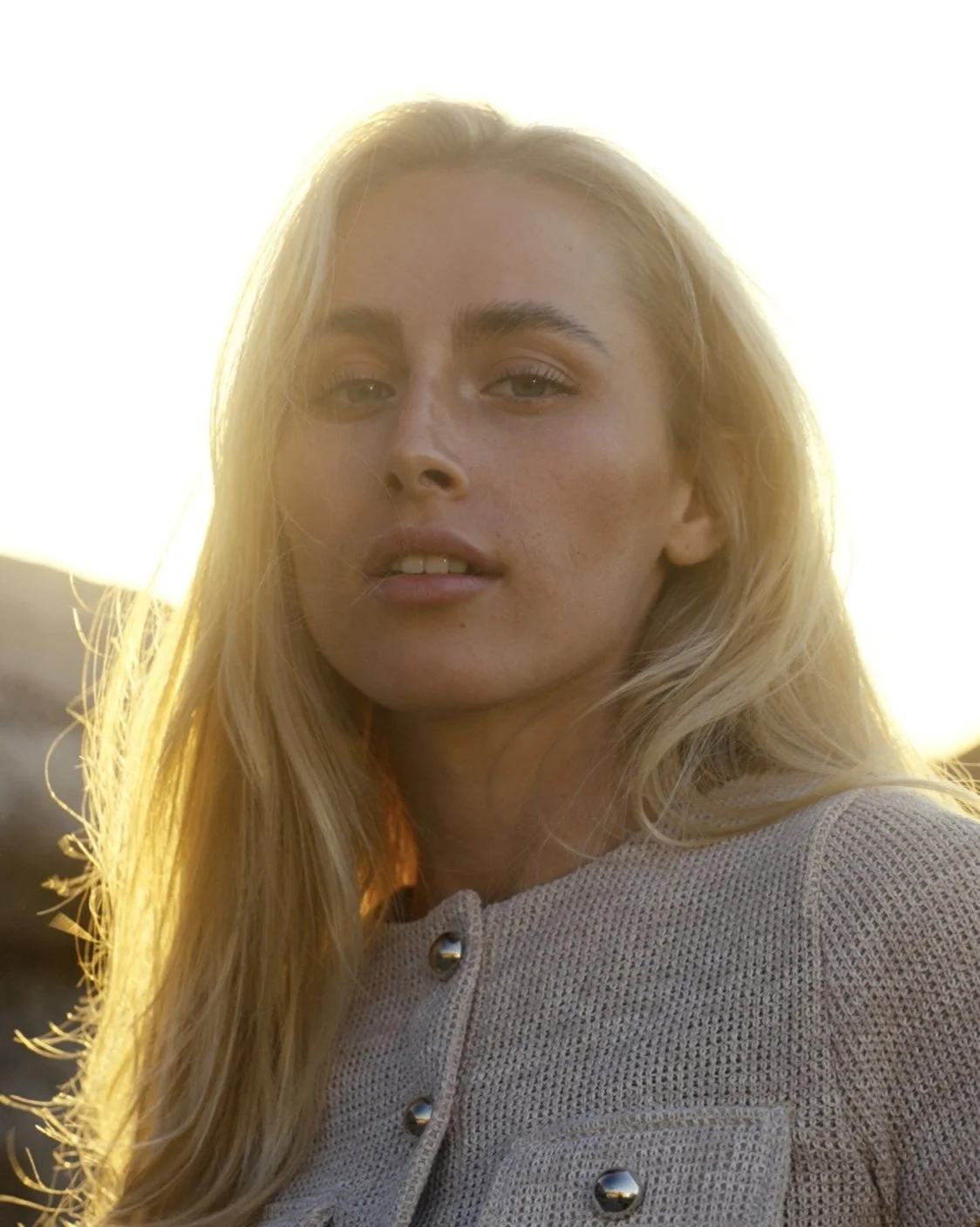 A woman with long blonde hair and light makeup looking at the camera, backlit by sunlight creating a warm glow.
