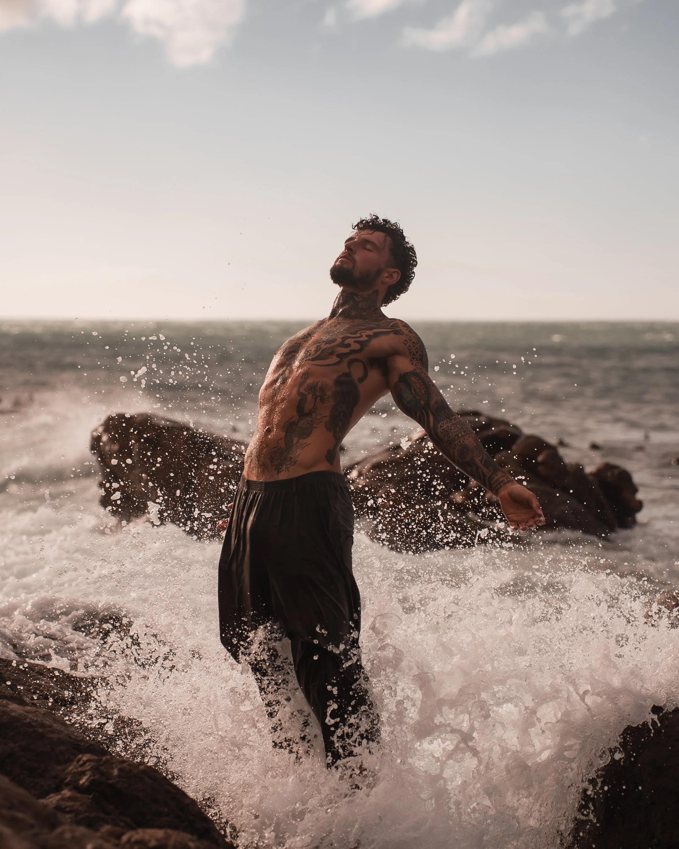 A shirtless man with tattoos standing in the ocean waves with rocks in the background, enjoying the water.