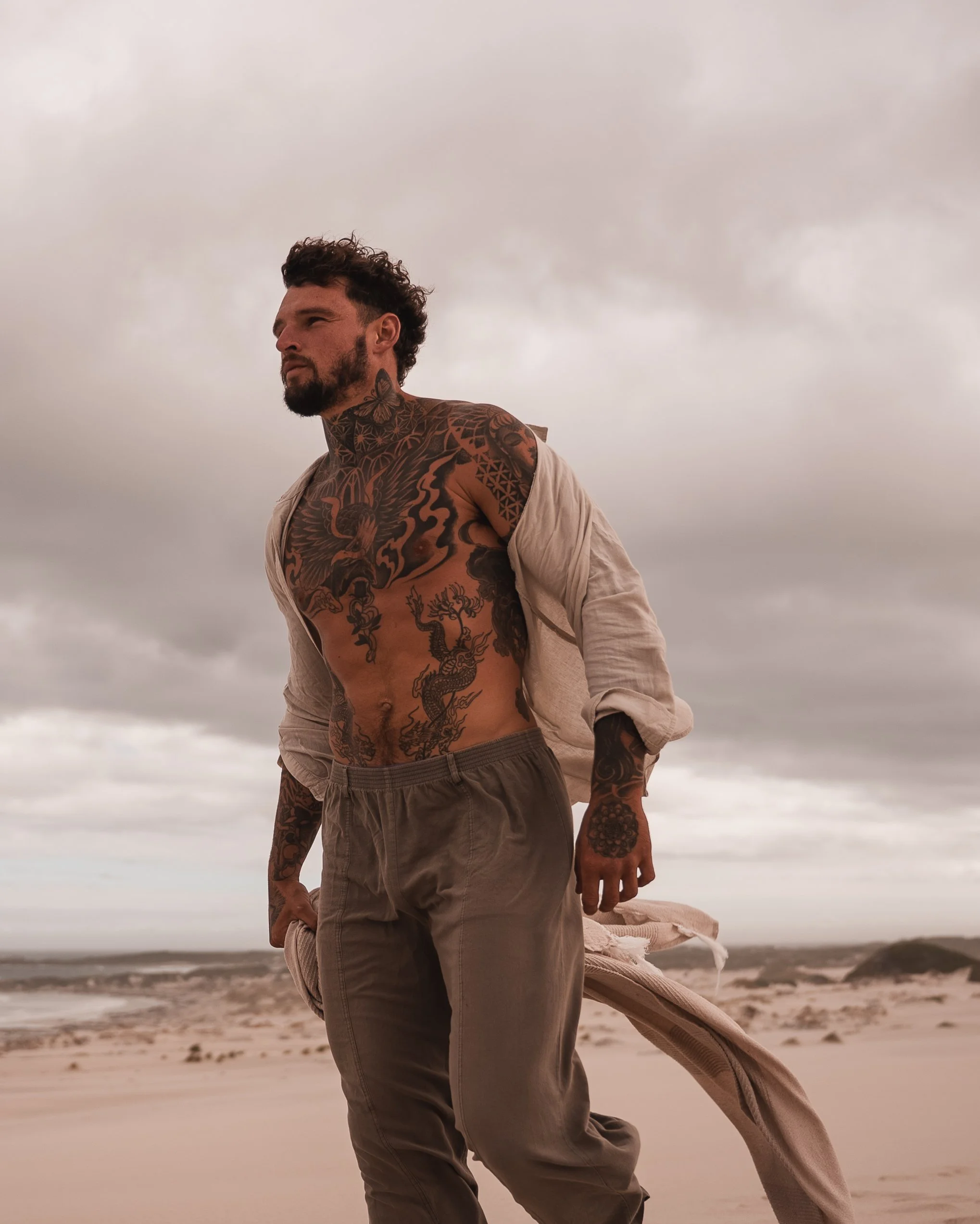 A tattooed man standing on a sandy beach with cloudy sky in the background.