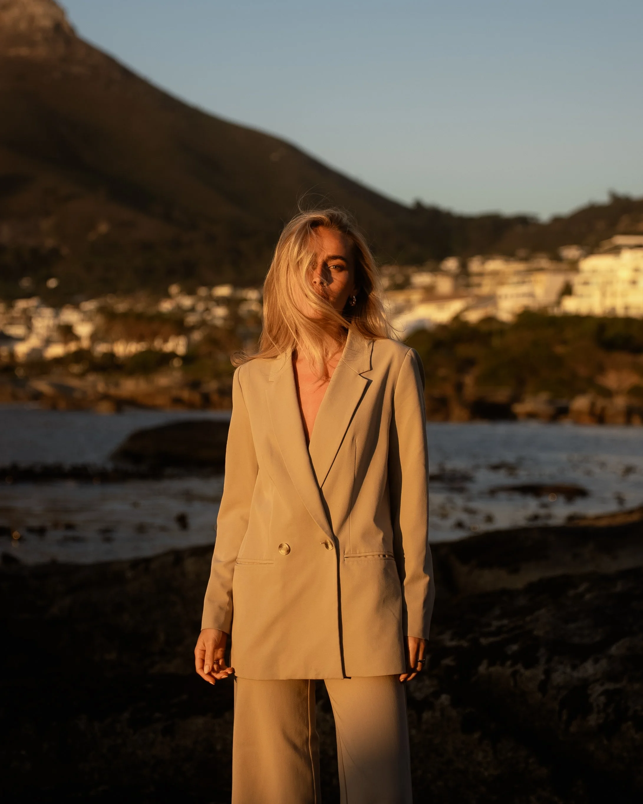 A woman in a beige suit standing outdoors at sunset with mountains and water in the background.