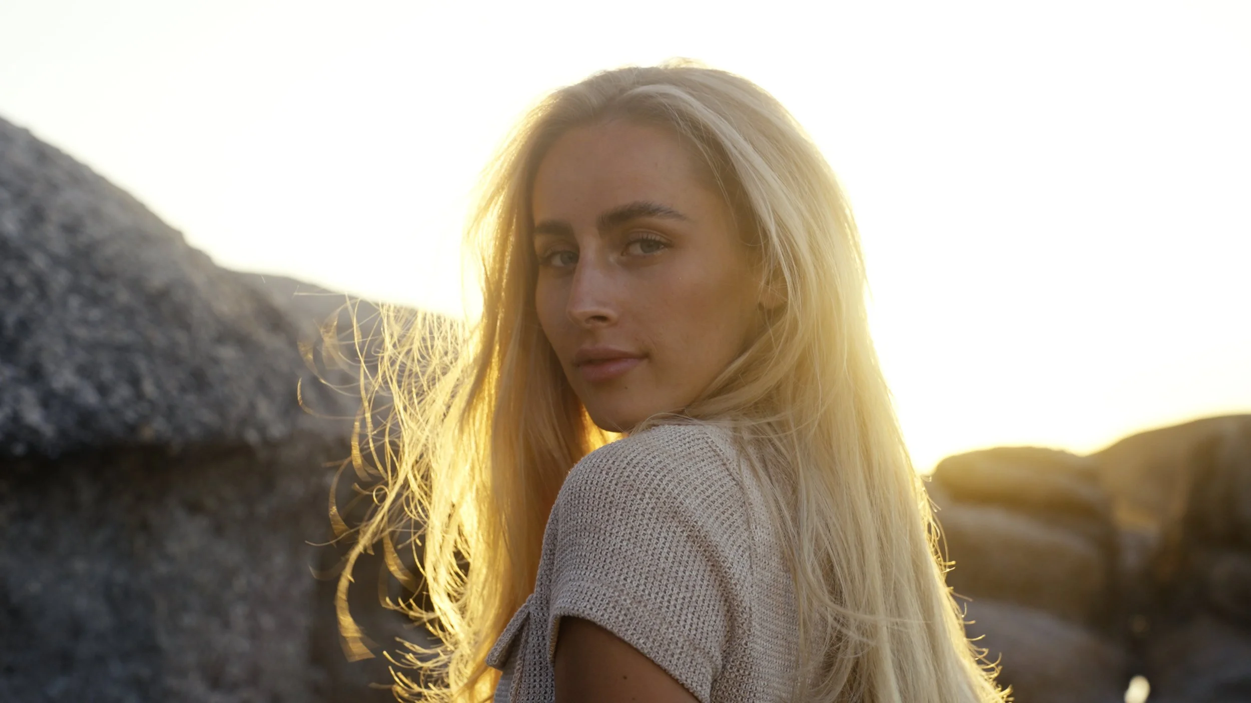 A young woman with long blonde hair looking over her shoulder at the camera, outdoors with rocks and sunlight in the background.