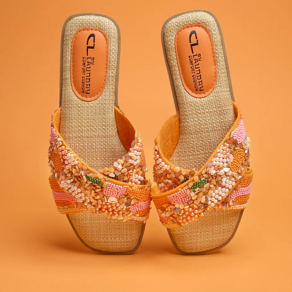 Pair of beige slip-on sandals with embellished straps featuring colorful beads, shells, and pearls, placed against an orange background.