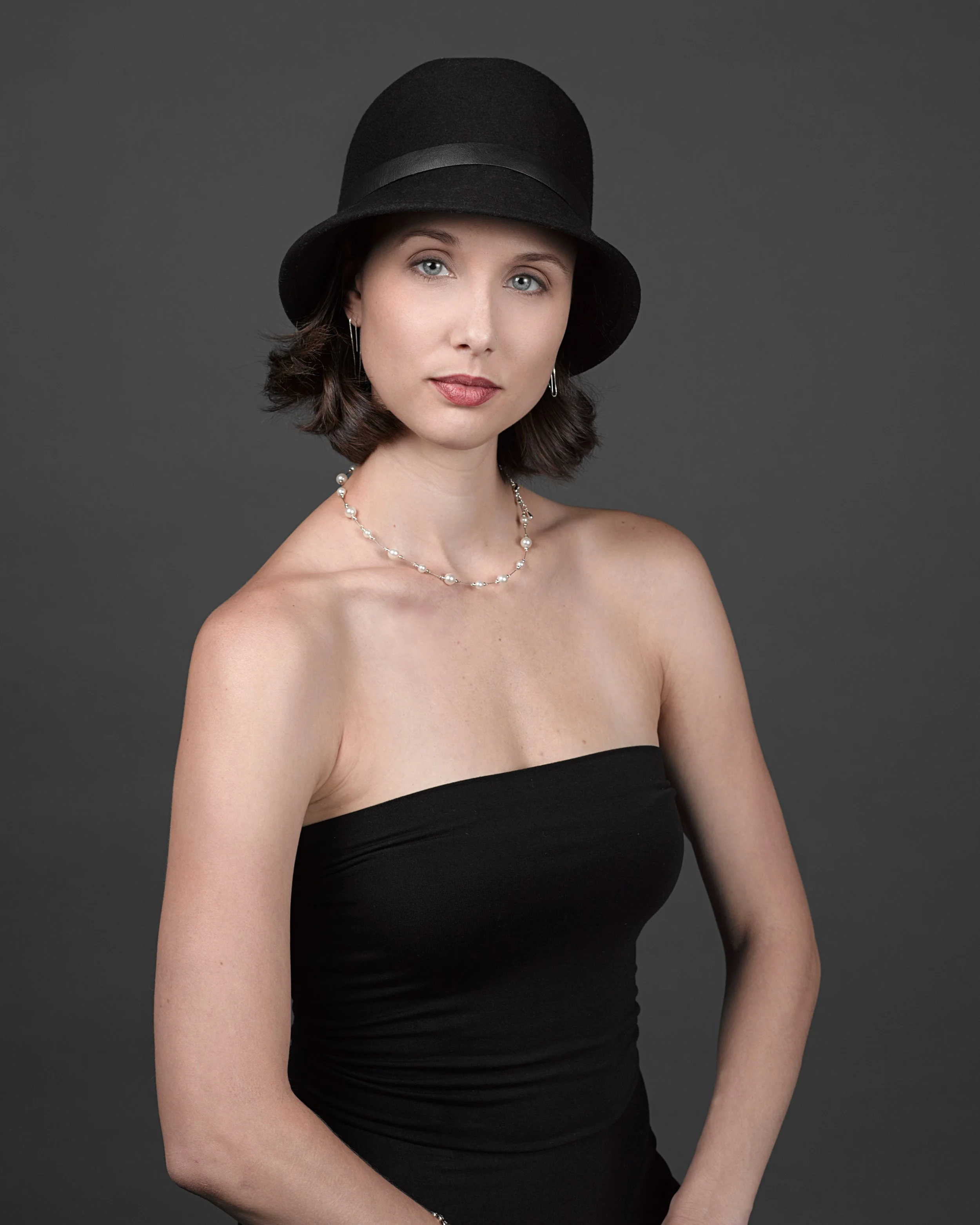 A woman with shoulder-length brown hair wearing a black strapless dress, a black hat, a pearl necklace, and matching earrings, posing against a dark gray background.