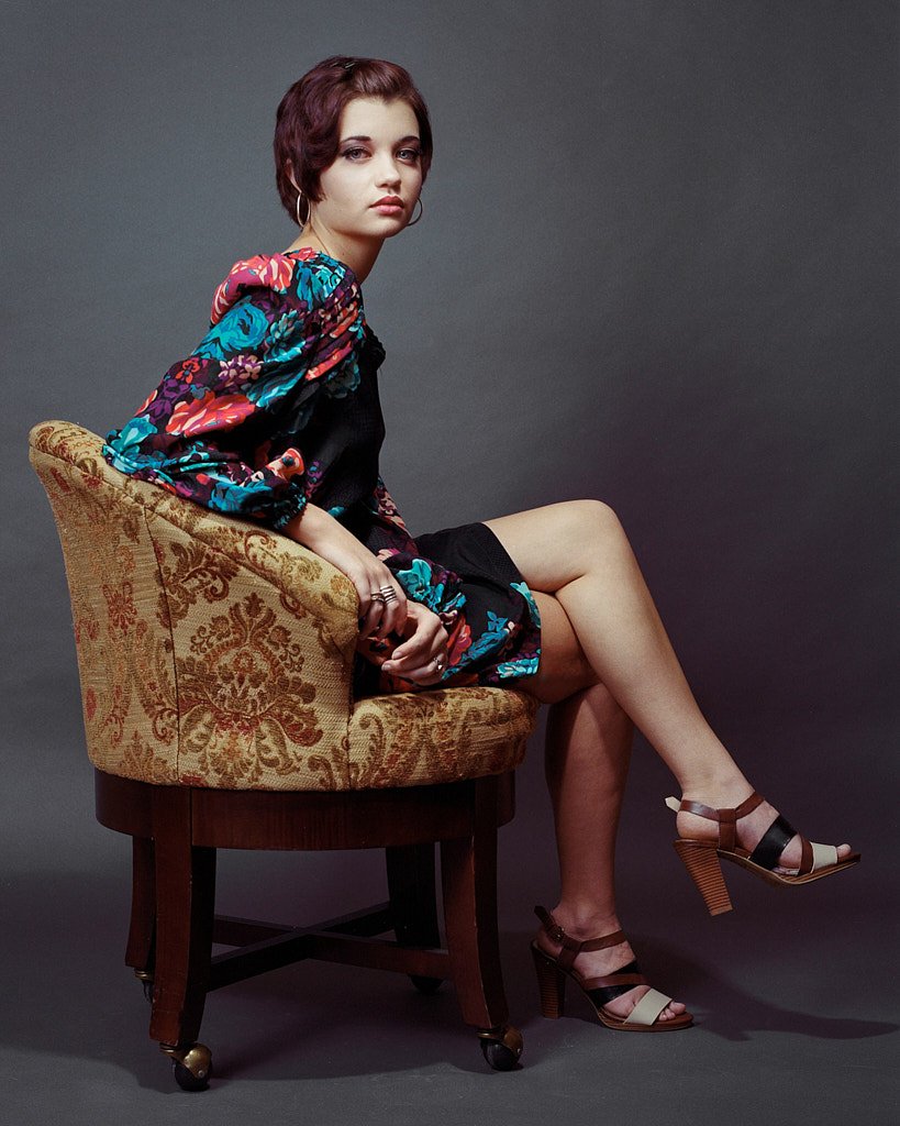 A young woman with short reddish-brown hair sitting sideways in an ornate vintage-style chair with floral upholstery, wearing a colorful, floral-patterned jacket and black shorts, with beige and brown high-heeled sandals, against a plain gray backgro