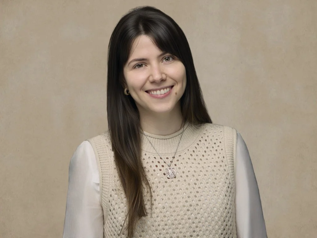 Professional headshot young woman with long brown hair, smiling, wearing a beige knit vest over a white blouse, with a silver necklace and earrings, standing against a plain beige background.