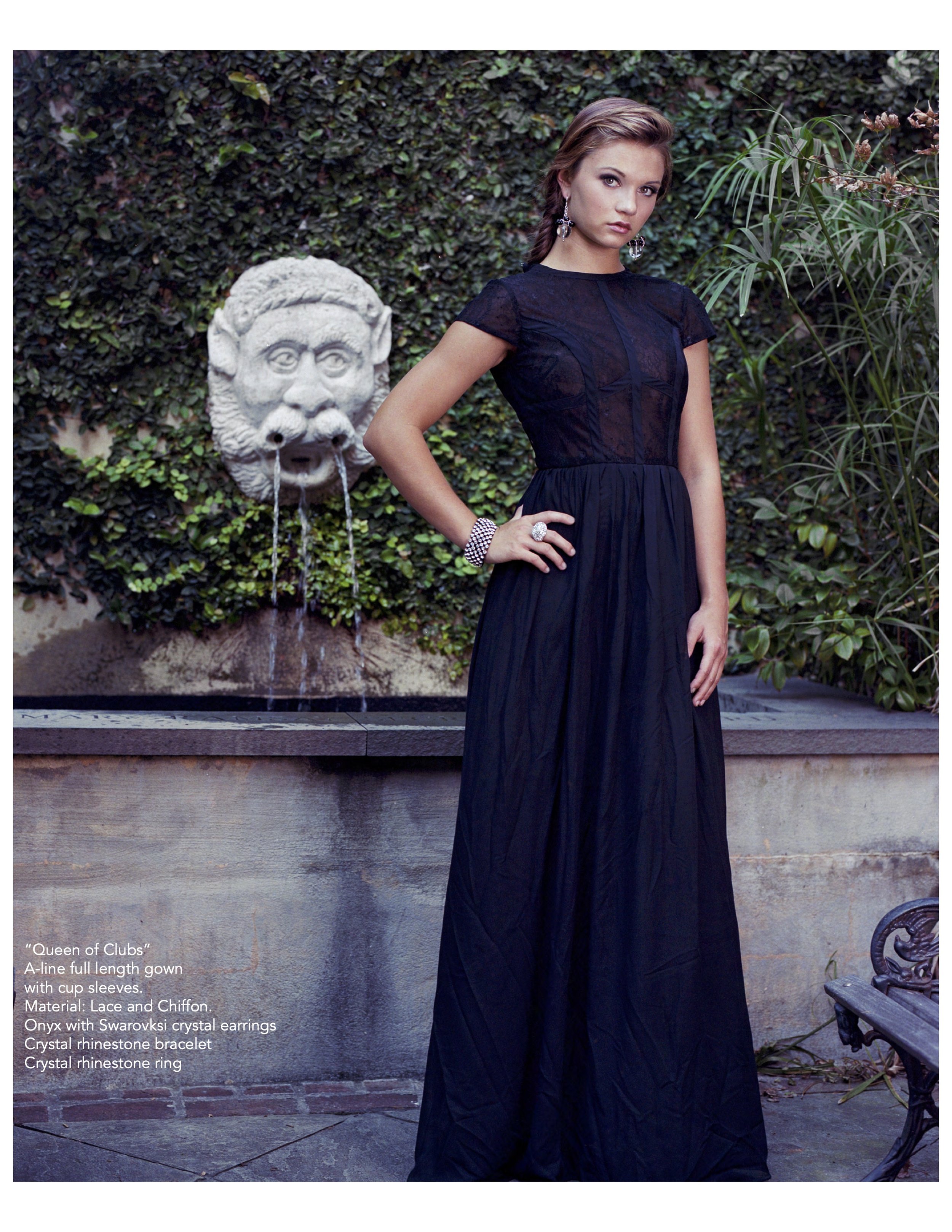 A young woman in a black full-length gown with cup sleeves standing outdoors in front of a fountain with a lion head spout, surrounded by greenery.