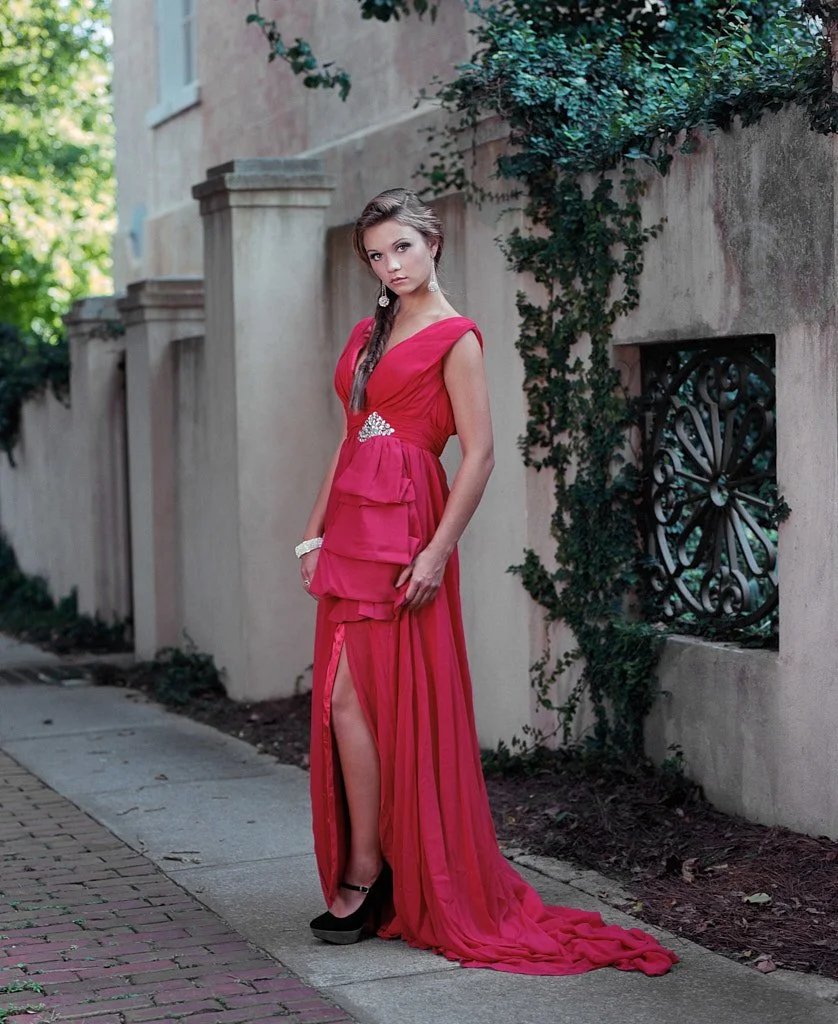 Young woman in a long, elegant pink dress with a slit, standing on a sidewalk next to a stucco wall with green ivy. She has long brown hair, wears earrings, a bracelet, and black high-heeled shoes.