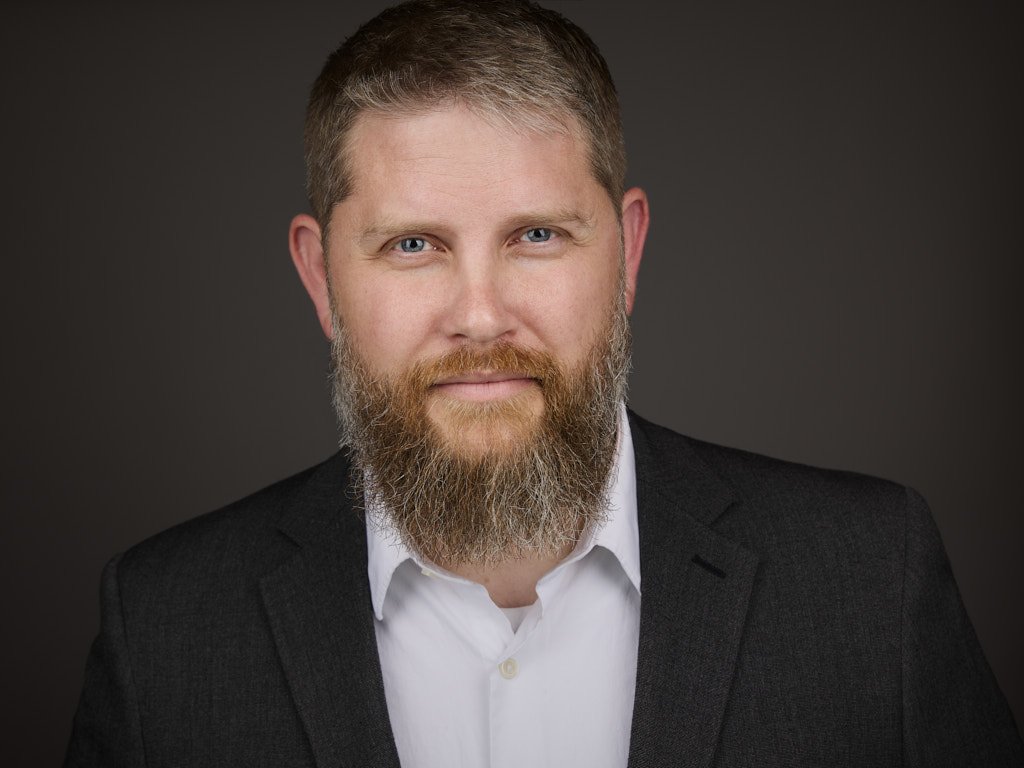 Portrait of a man with short hair and a full beard, wearing a white shirt and dark blazer, against a dark background.
