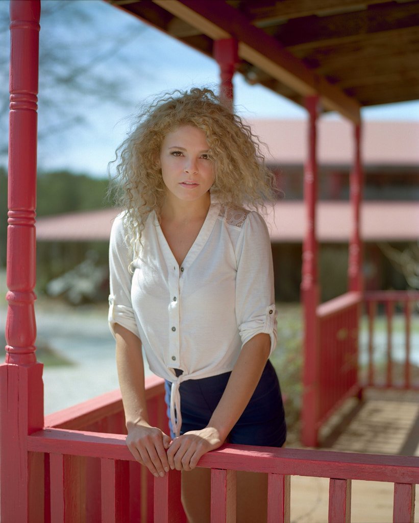 Young woman with curly blonde hair leaning on a red wooden railing outside