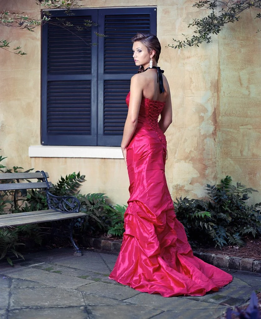 A woman in a red strapless gown with a ruffled train standing outdoors near a bench and foliage, with closed blue shutters on a yellow wall in the background.