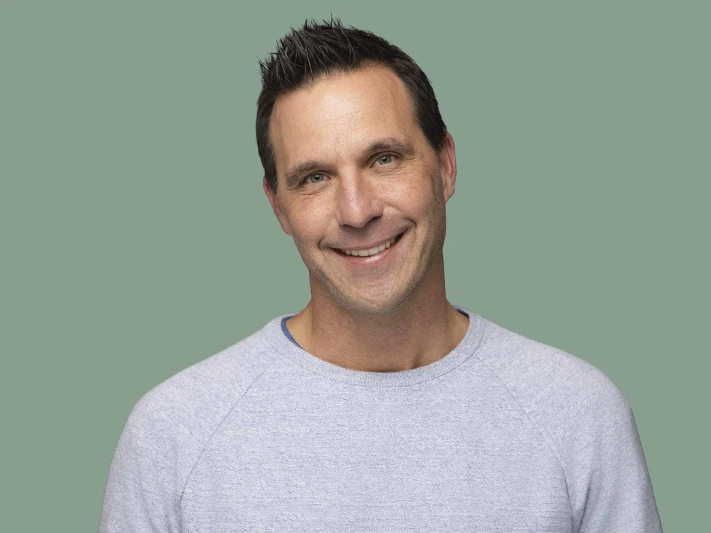 A man with short, dark hair, smiling, wearing a light gray sweatshirt, against a solid green background.