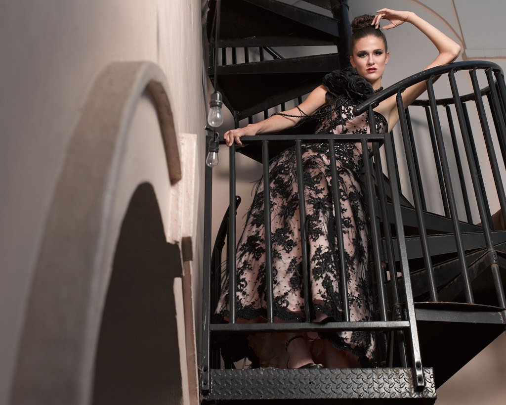 A woman in a black lace dress poses on a spiral staircase, leaning with one arm on the railing and the other arm raised above her head.