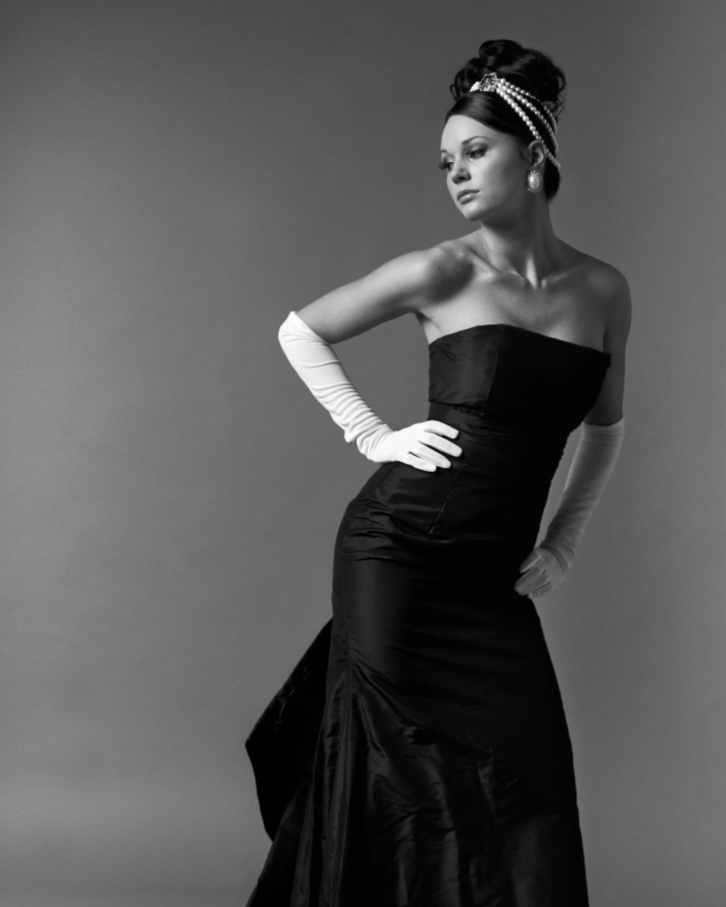 A woman dressed in a strapless black gown with long white gloves, her hair styled in an elaborate updo with pearl accessories, standing against a plain background.